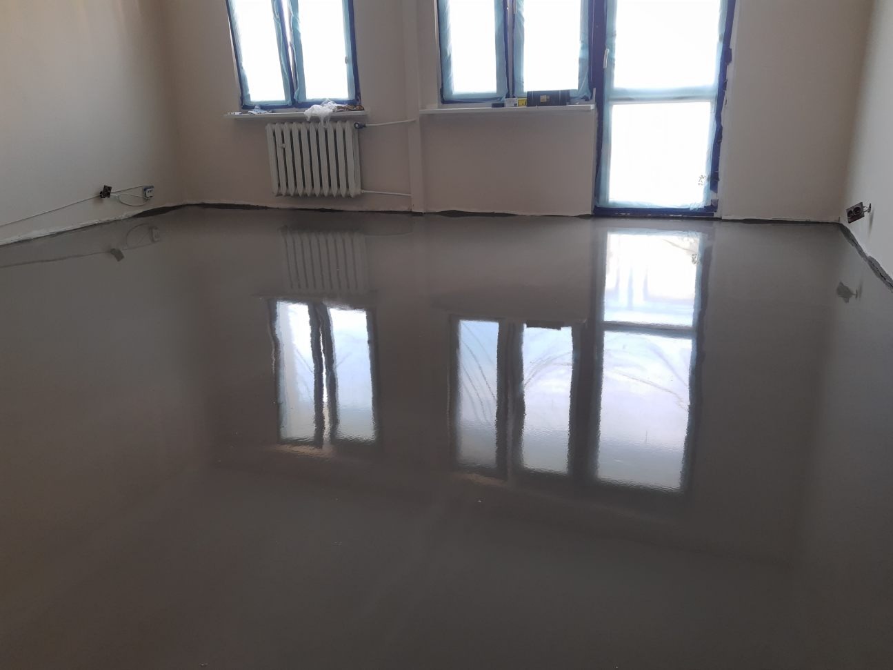 Świeżo wylana, samopoziomująca wylewka podłogowa w pokoju, odbijająca okna i kaloryfer, z widocznymi zabezpieczeniami okien folią.