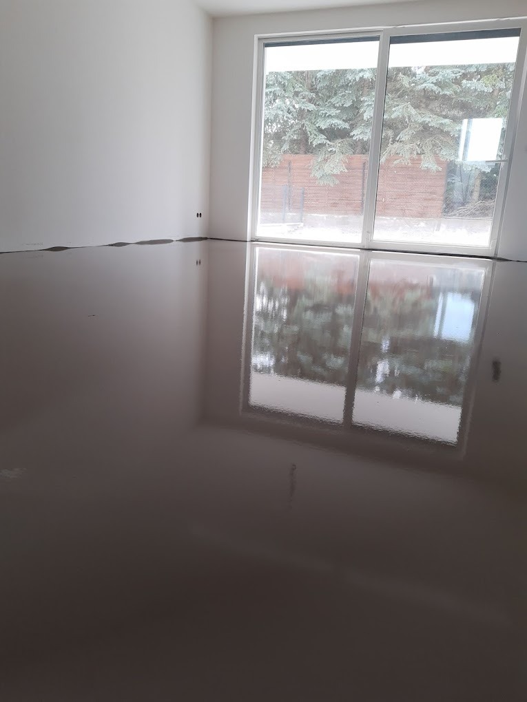 Gładka, szara posadzka w minimalistycznym wnętrzu z dużym oknem przesuwnym, odbijającym zieleń drzew na zewnątrz.