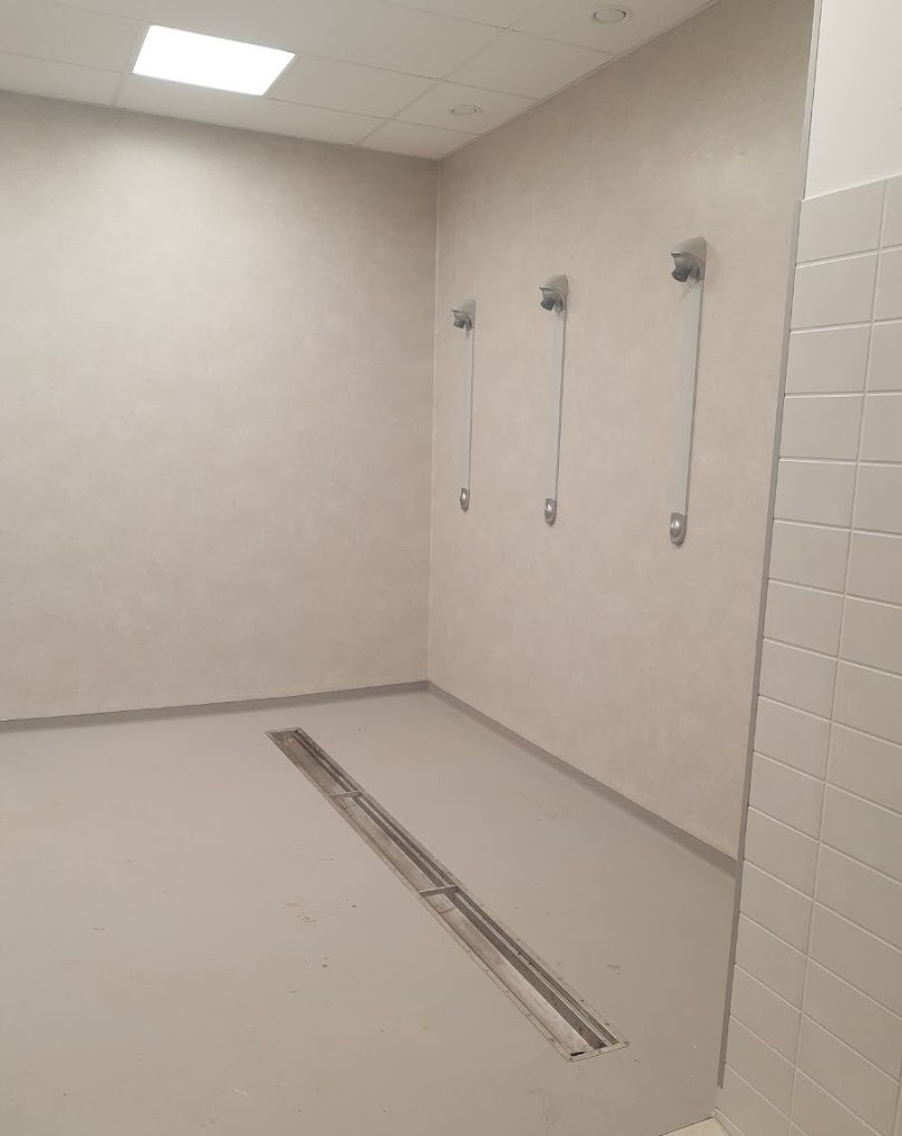 Jasnoszara, jednolita wykładzina podłogowa w pomieszczeniu z odpływem liniowym i trzema wieszakami na ścianie.