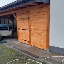 Usługi budowlane MarBud Mariusz Olszewski - Drewniana brama wjazdowa z czarnymi zawiasami, przylegająca do białej ściany budynku. Na podłożu widoczna kostka brukowa i fragment węża ogrodowego.