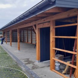 Usługi budowlane MarBud Mariusz Olszewski - Drewniana wiata garażowa z ciemnym dachem i rynnami, widok z zewnątrz. W tle osoba wykonująca prace wykończeniowe. Kostka brukowa i trawnik przed wiatą.