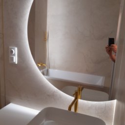 Usługi budowlane MarBud Mariusz Olszewski - Nowoczesna łazienka z podświetlanym lustrem w kształcie półksiężyca, białą umywalką nablatową i złotą armaturą, odbicie osoby robiącej zdjęcie w lustrze.