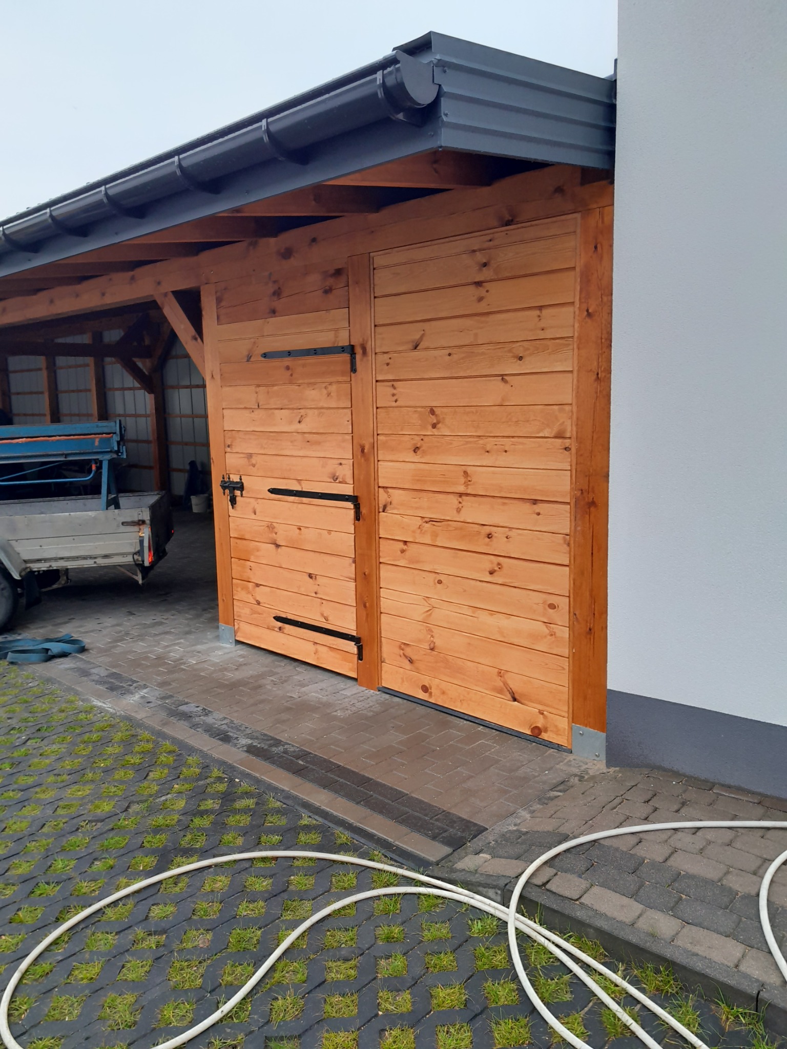 Drewniana brama wjazdowa z czarnymi zawiasami, przylegająca do białej ściany budynku. Na podłożu widoczna kostka brukowa i fragment węża ogrodowego.