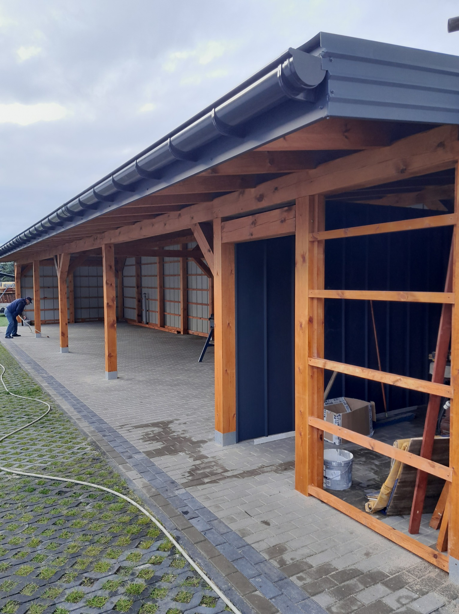 Drewniana wiata garażowa z ciemnym dachem i rynnami, widok z zewnątrz. W tle osoba wykonująca prace wykończeniowe. Kostka brukowa i trawnik przed wiatą.