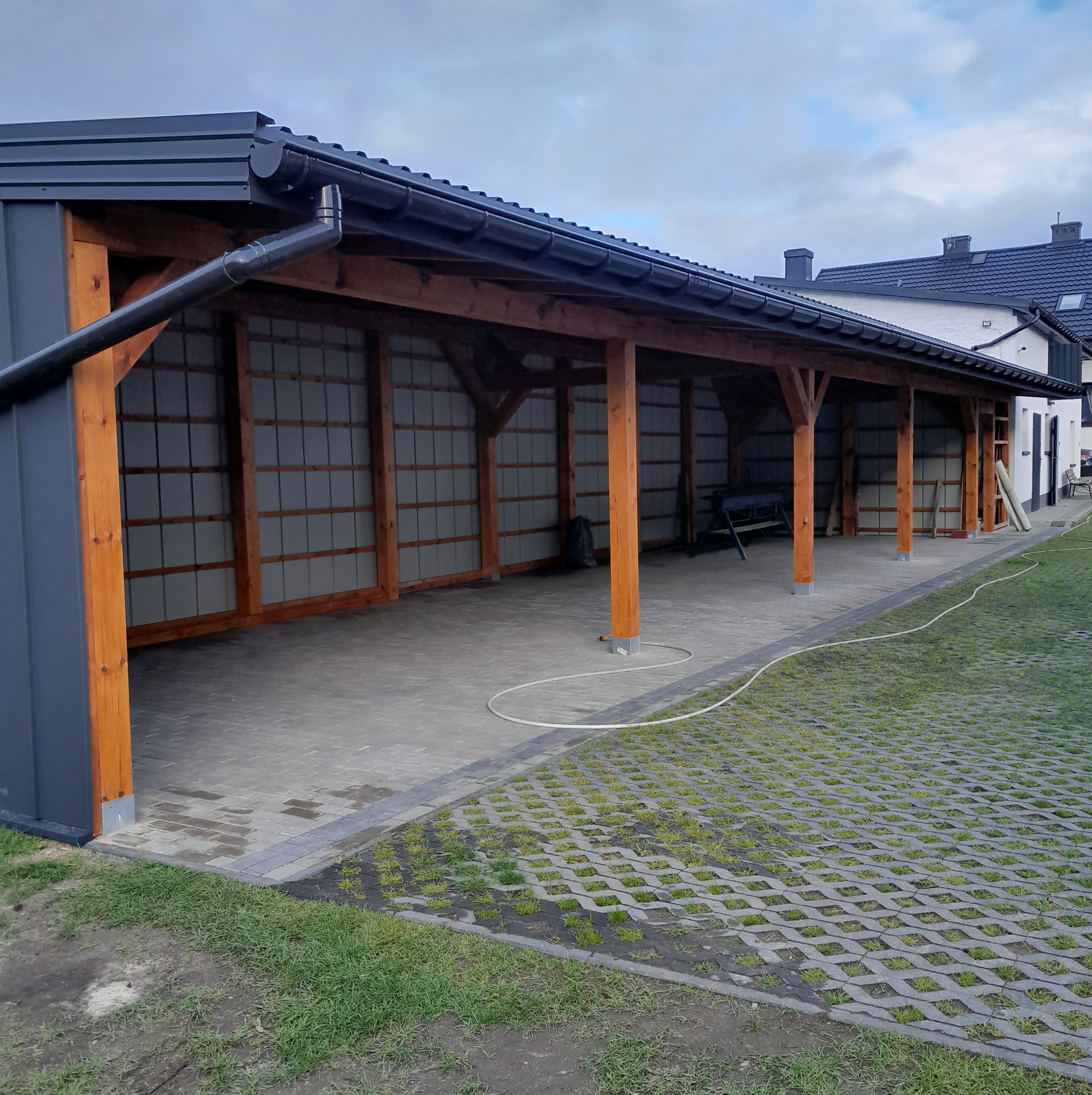 Zadaszenie z drewnianą konstrukcją i szarą blachą trapezową, widok z zewnątrz na kostkę ażurową i budynek w tle, pochmurne niebo.