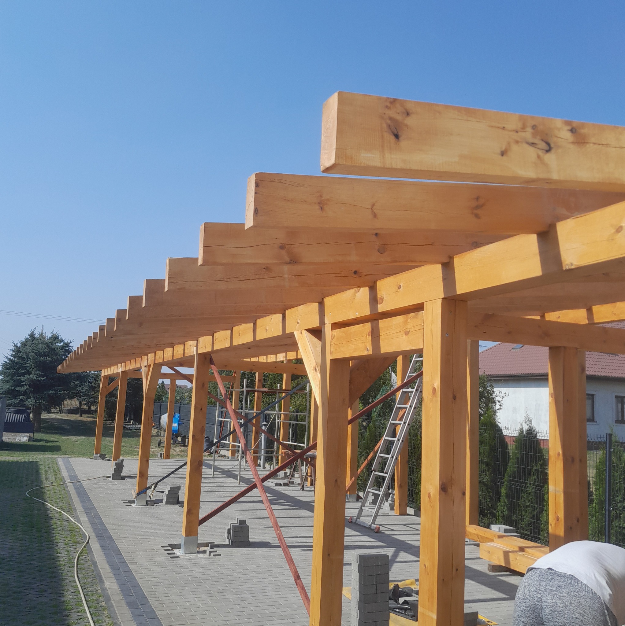 Drewniana konstrukcja z belek, prawdopodobnie zadaszenie tarasu w trakcie budowy. Widoczne rusztowania i elementy montażowe, jasne drewno na tle błękitnego nieba.