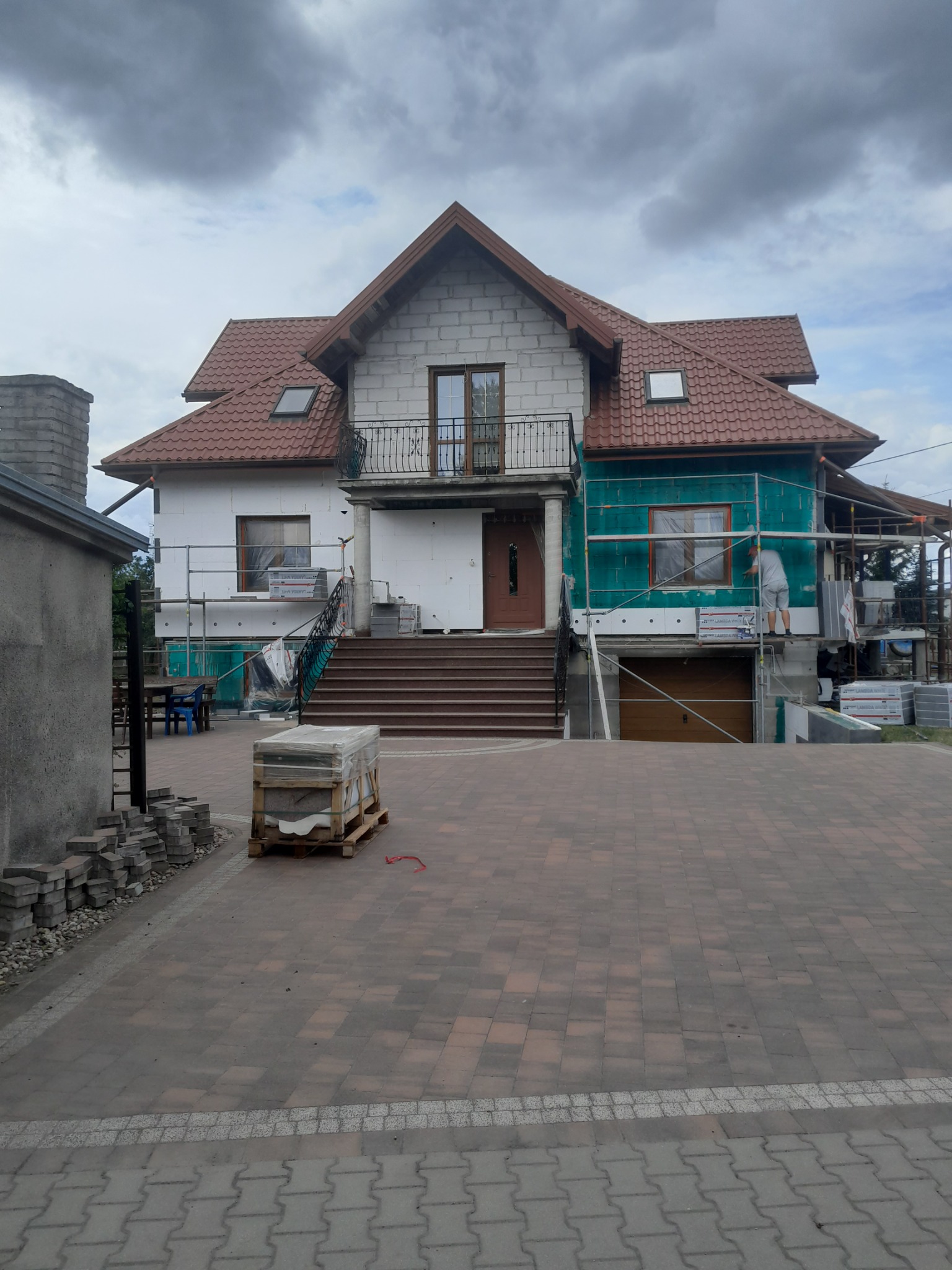 Dom w trakcie ocieplania: styropian na ścianach, rusztowania. Część elewacji z białym styropianem, część z zieloną folią. Na pierwszym planie kostka brukowa.