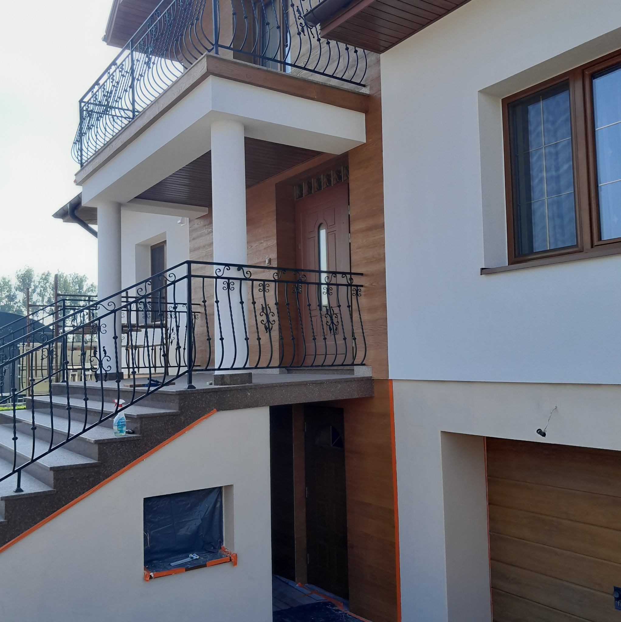 Elewacja domu z balkonami i schodami, wykończona w jasnych kolorach z elementami drewna. Widoczne detale architektoniczne i balustrady. Częściowo zakryte okno folią.