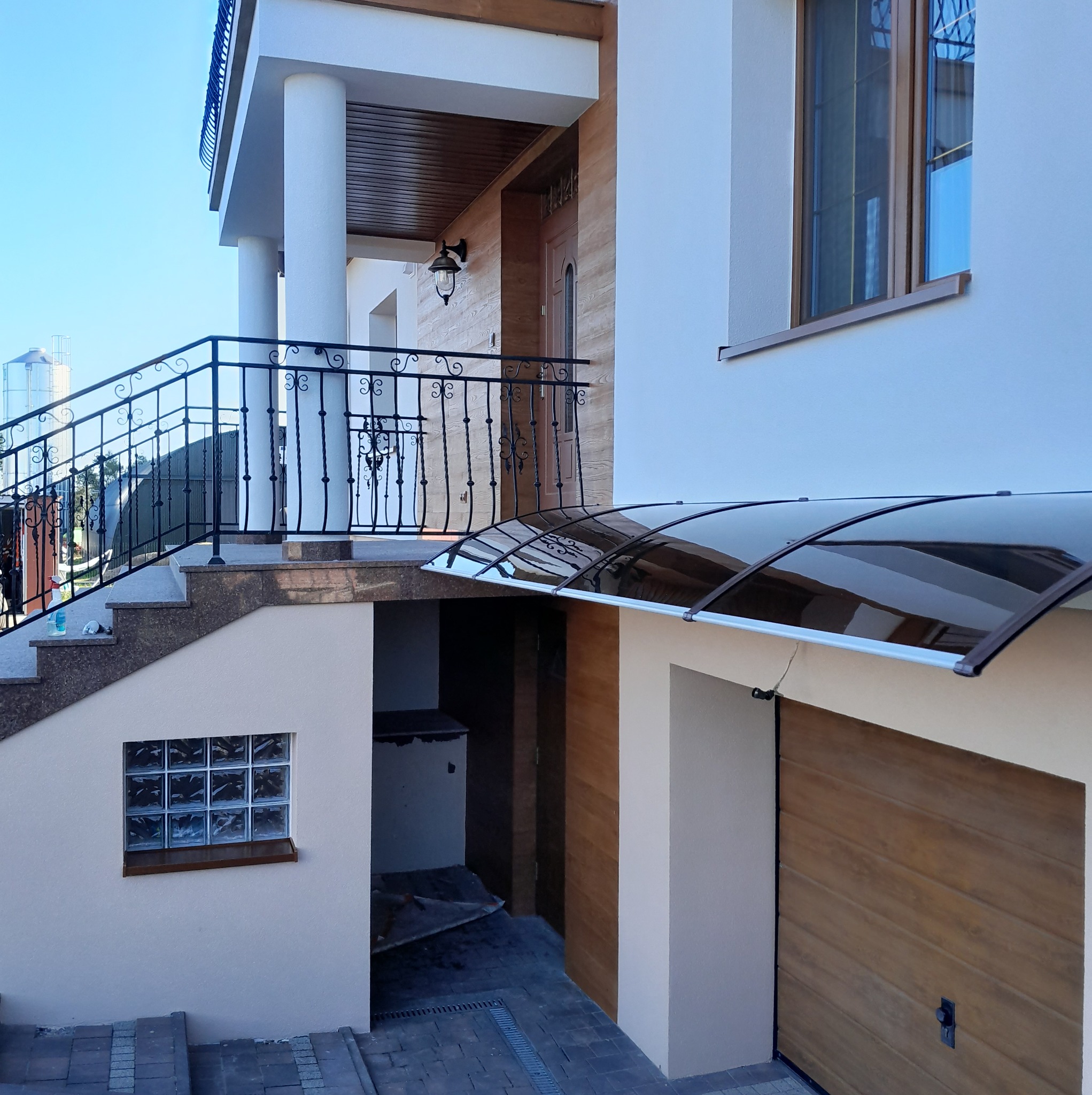 Elewacja domu z zadaszeniem nad garażem, widoczne schody z ozdobną balustradą i okno z luksferów. Jasna kolorystyka budynku i brązowe akcenty.