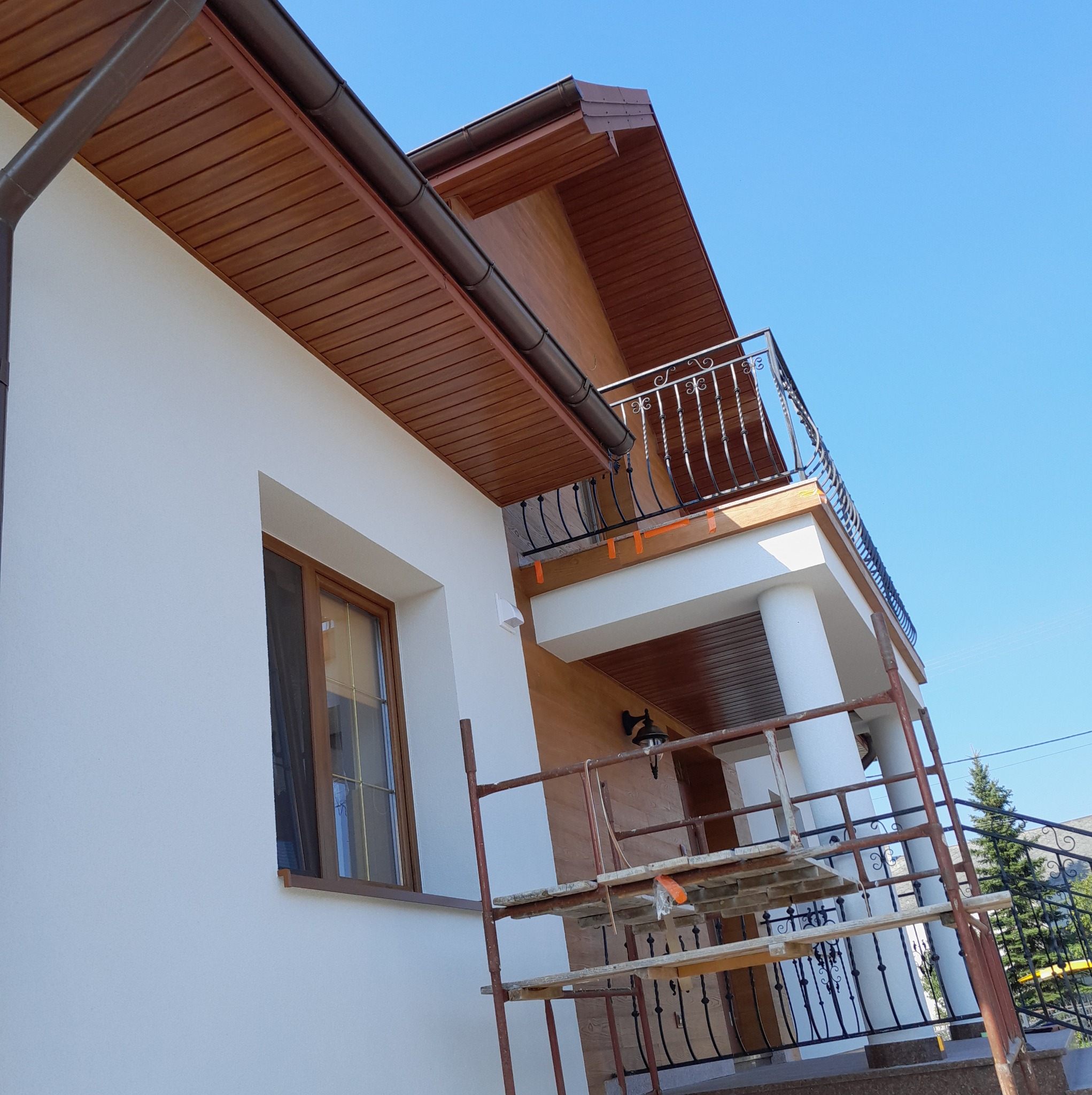 Elewacja domu z balkonem i rusztowaniem, białe ściany i brązowe elementy wykończeniowe, widok z dołu.