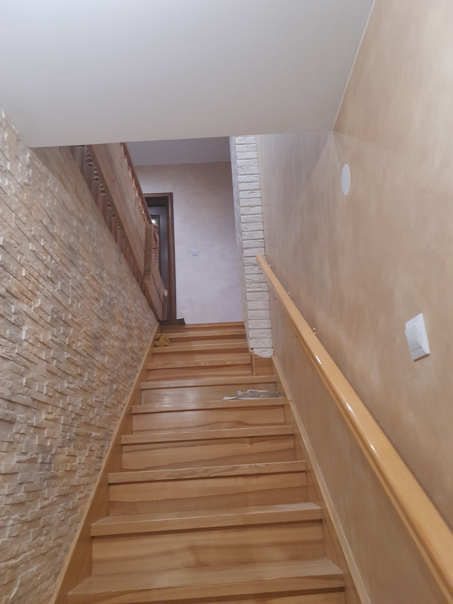Klatka schodowa z drewnianymi schodami, ściana wyłożona imitacją kamienia, druga ściana w odcieniach beżu, drewniana balustrada.