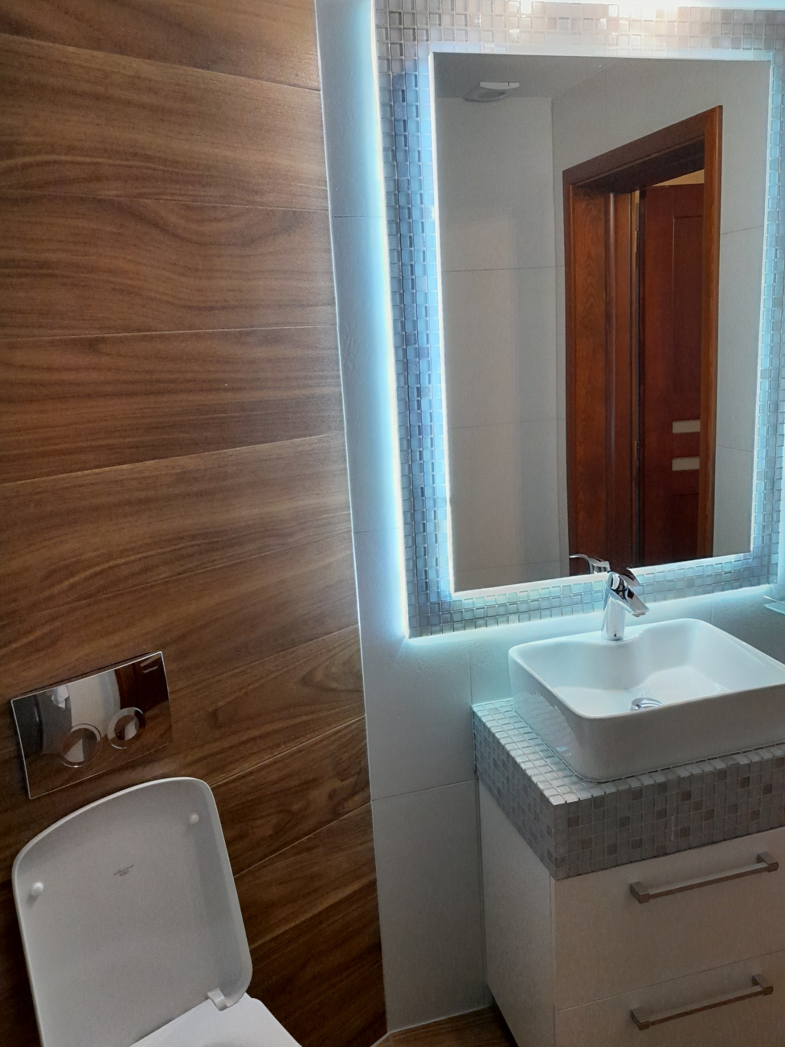 Nowoczesna łazienka w Konopkach z drewnopodobnymi płytkami na ścianie, podświetlanym lustrem mozaikowym, białą umywalką nablatową i wiszącą toaletą.