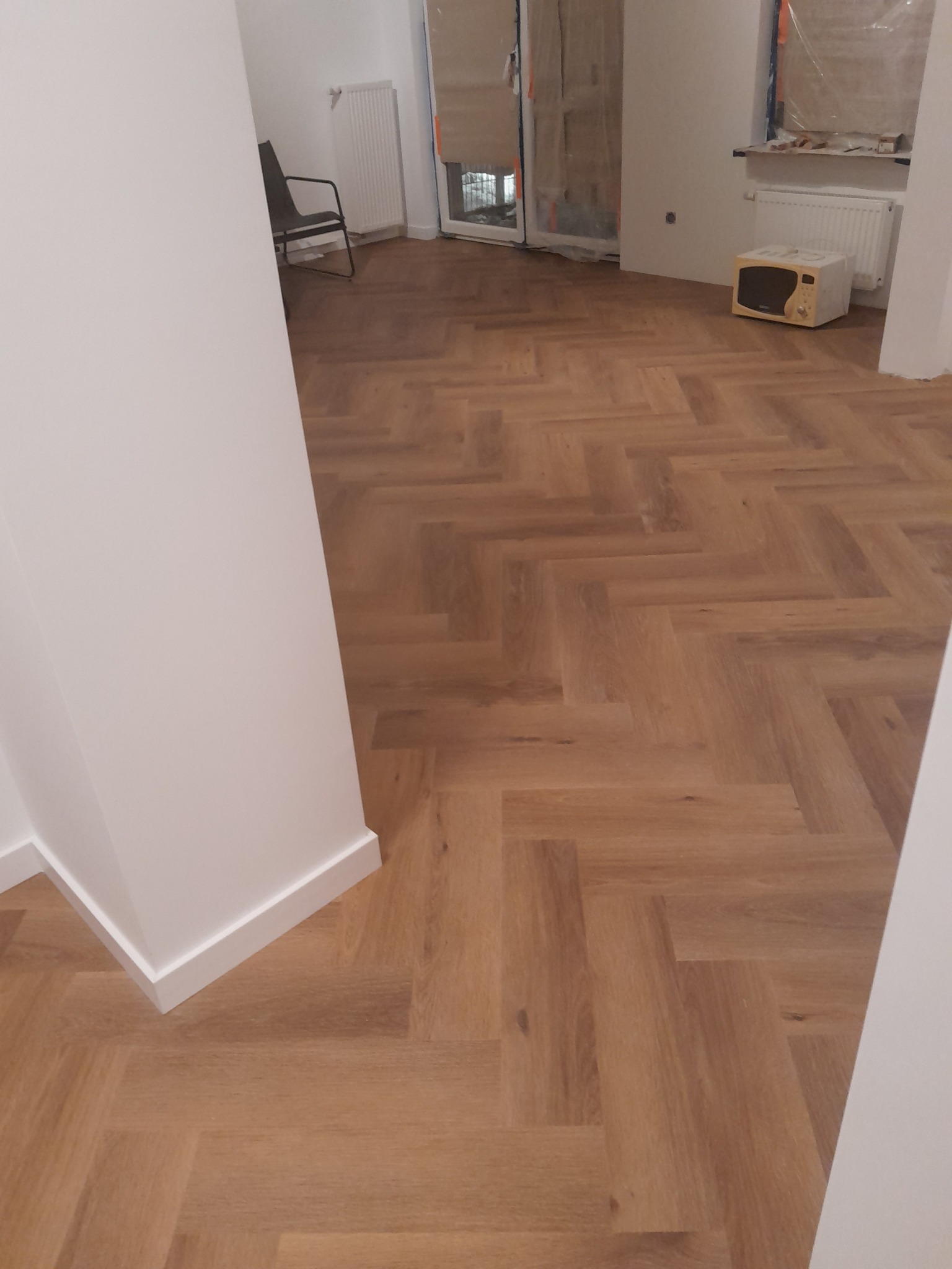 Ułożona podłoga z paneli w jodełkę, widok z perspektywy wejścia do pomieszczenia, białe ściany z listwami przypodłogowymi, w tle krzesło i mikrofalówka w remonowanym pokoju.