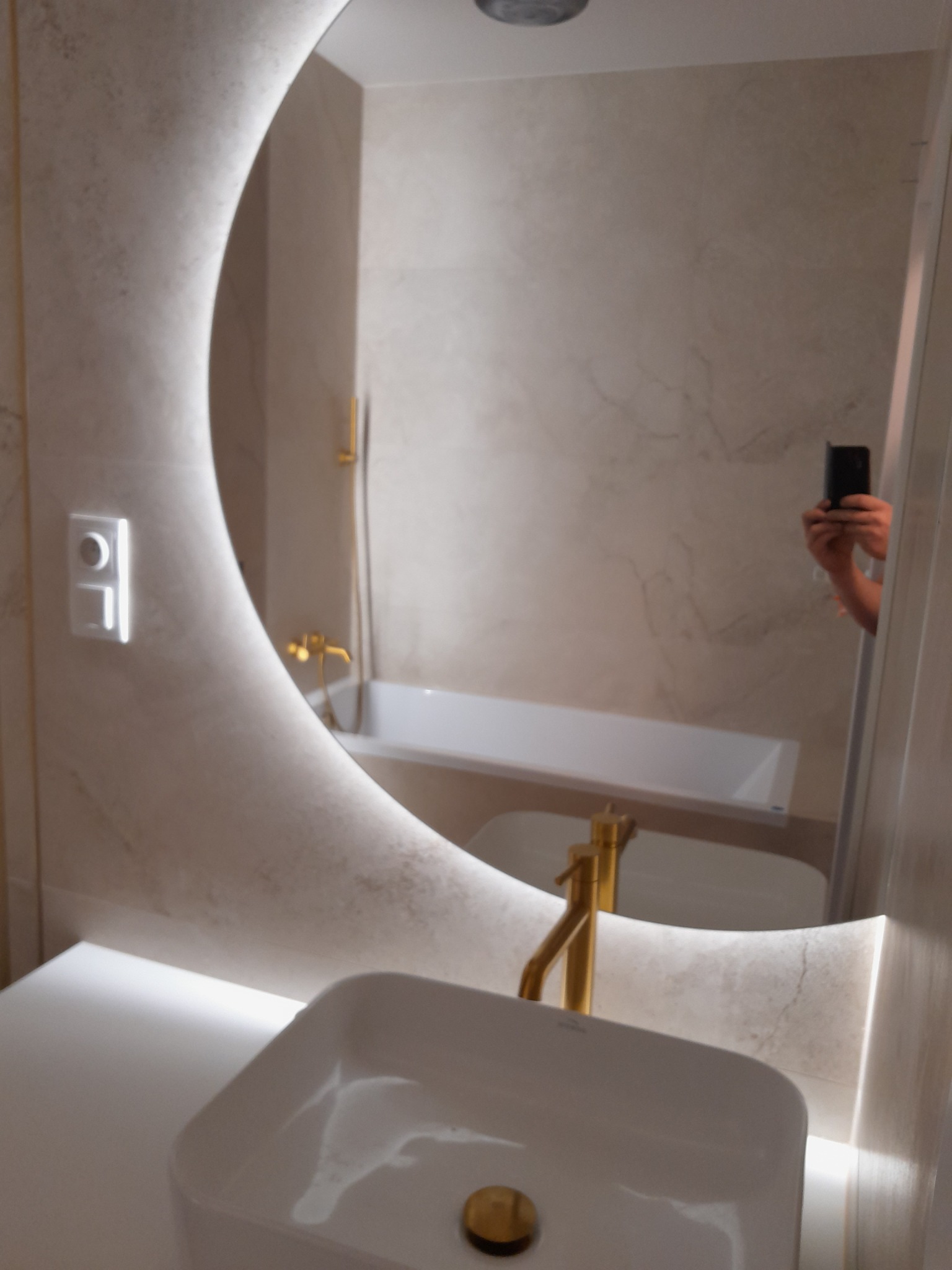 Nowoczesna łazienka z podświetlanym lustrem w kształcie półksiężyca, białą umywalką nablatową i złotą armaturą, odbicie osoby robiącej zdjęcie w lustrze.
