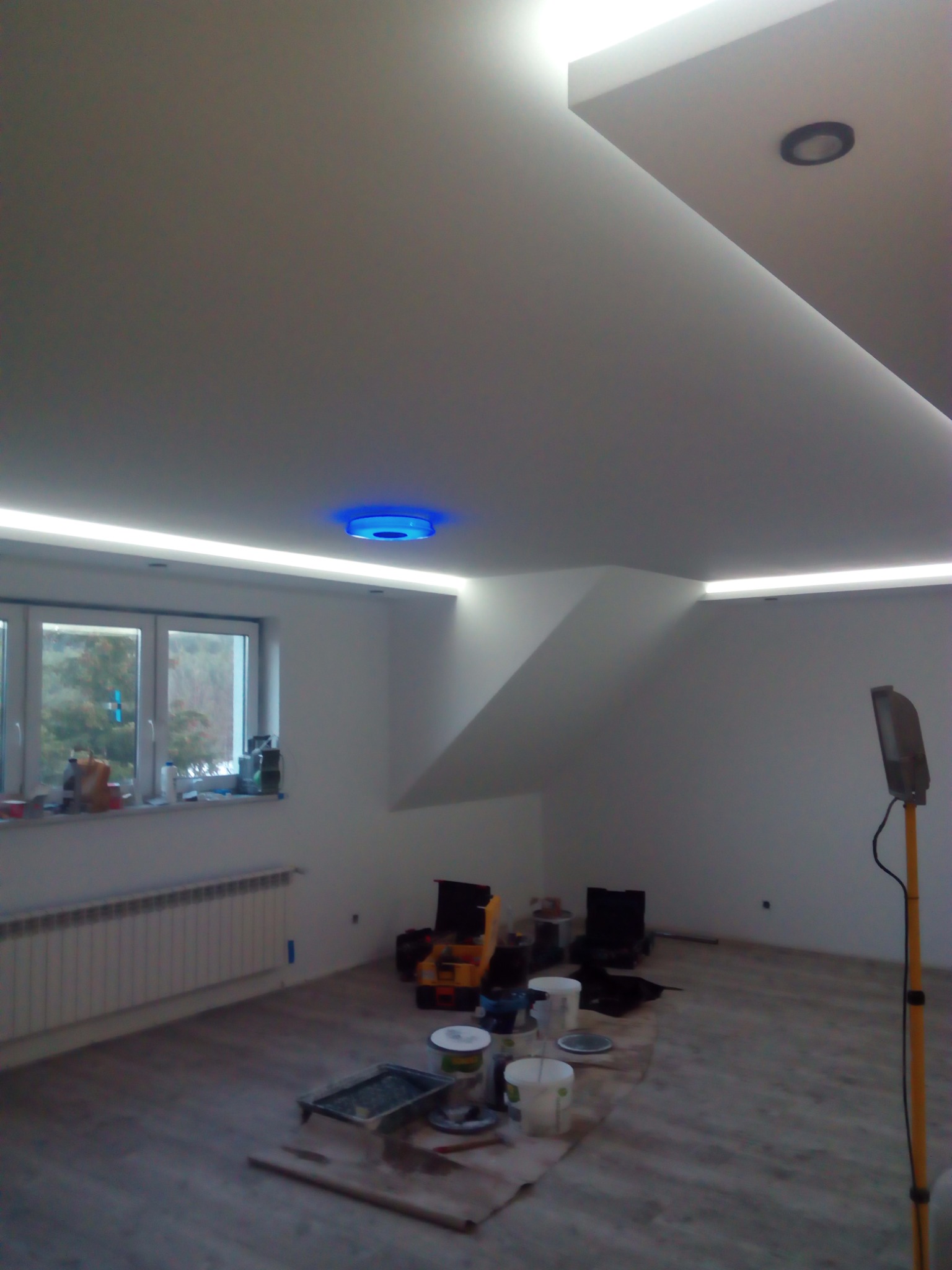 Pokój w trakcie prac wykończeniowych: białe ściany z listwami LED pod sufitem, okno z widokiem na zewnątrz, grzejnik, narzędzia malarskie i lampa oświetleniowa na statywie.