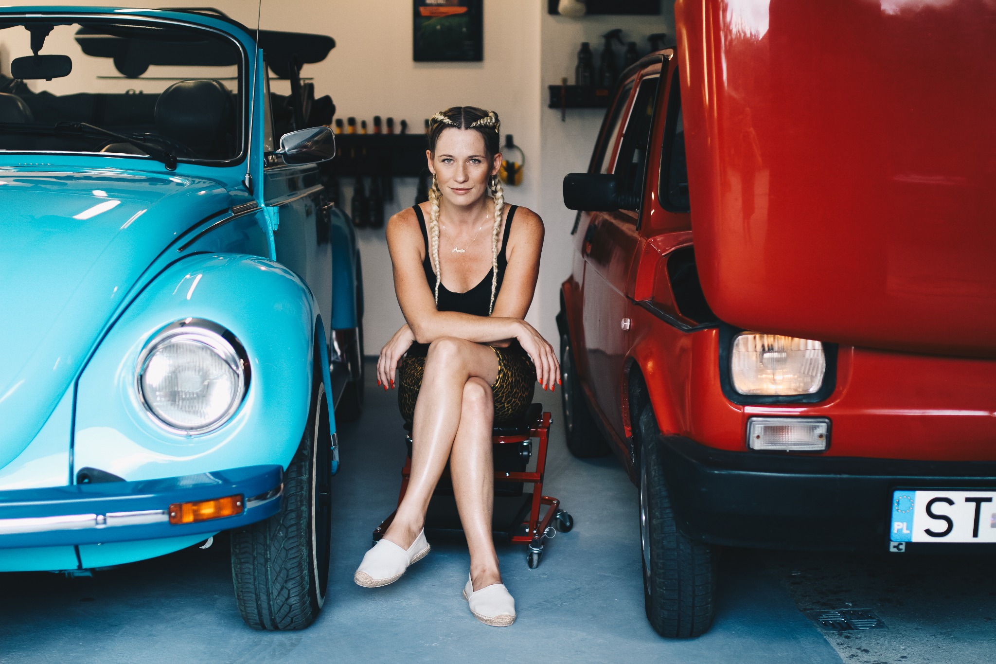 Kobieta siedzi na krześle warsztatowym pomiędzy turkusowym VW Garbusem kabrioletem a czerwonym Fiatem 126p z otwartą maską, w tle widoczne półki z preparatami samochodowymi.