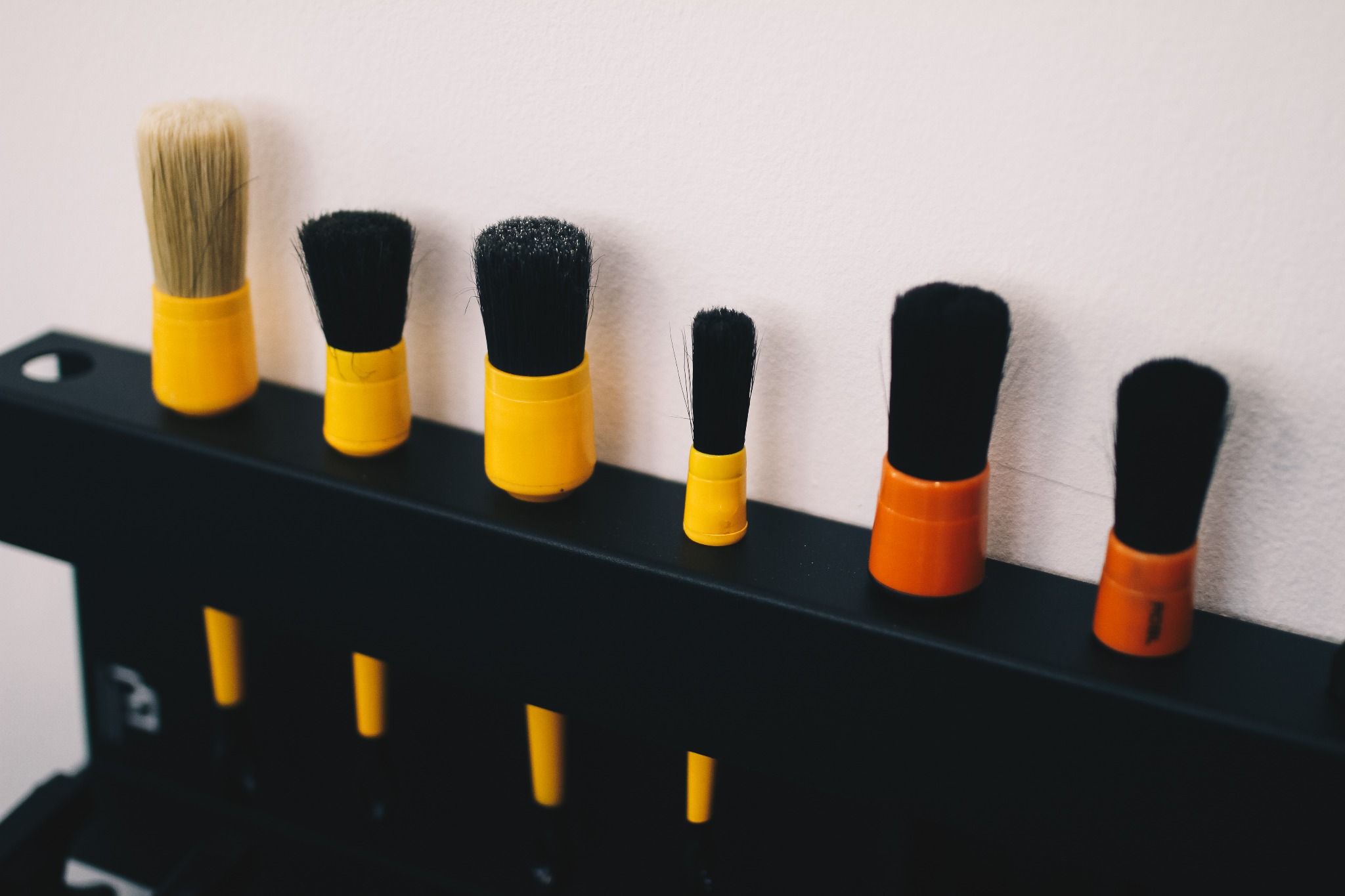 Rząd pędzli z czarnym włosiem i żółtymi (lub pomarańczowymi) uchwytami, umieszczonych w czarnym metalowym stojaku na tle jasnej ściany.