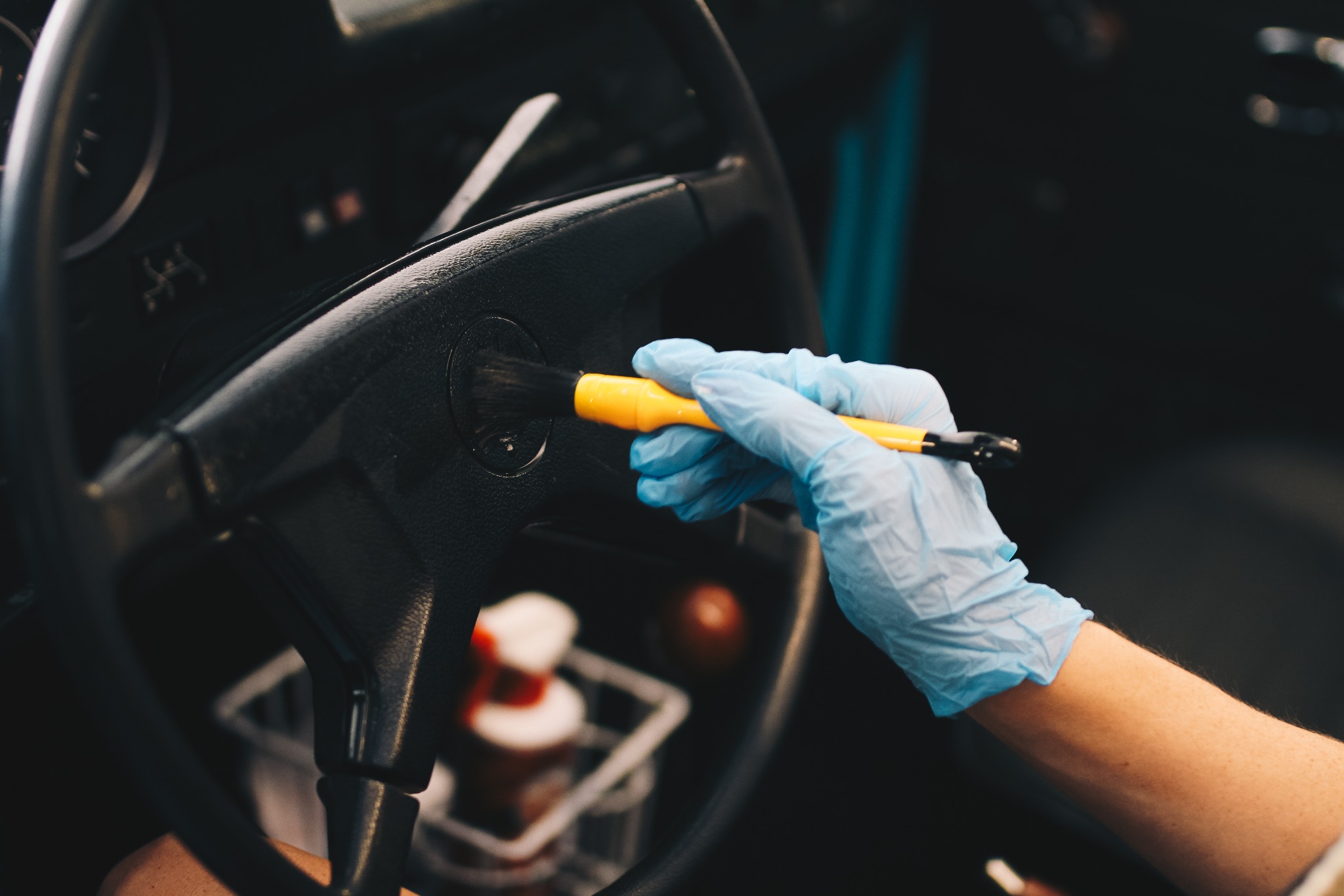 Czyszczenie kierownicy samochodu pędzlem z żółtą rączką przez osobę w niebieskiej rękawiczce, widoczna konsystencja materiału kierownicy i detale wnętrza pojazdu.