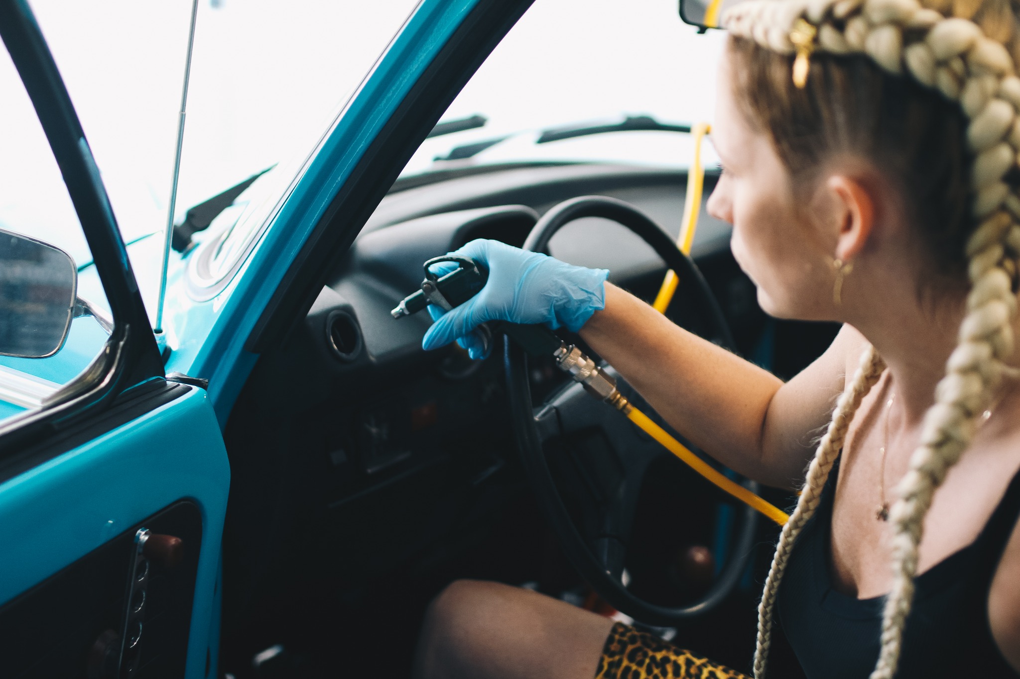 Kobieta w niebieskich rękawiczkach czyści wnętrze turkusowego samochodu za pomocą sprężonego powietrza, widoczny fragment deski rozdzielczej i kierownicy.