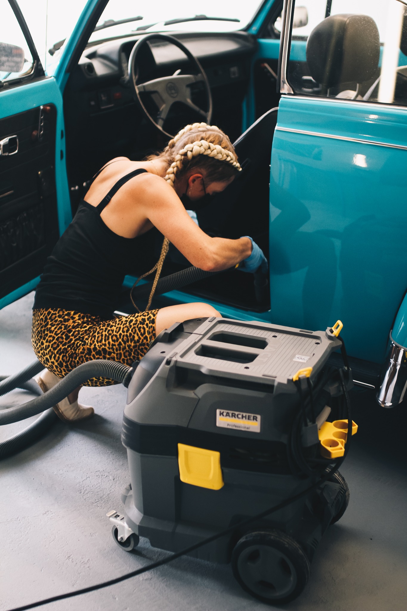 Kobieta w bluzie na ramiączkach i legginsach w panterkę czyści wnętrze turkusowego Volkswagena Garbusa przy użyciu odkurzacza Kärcher.