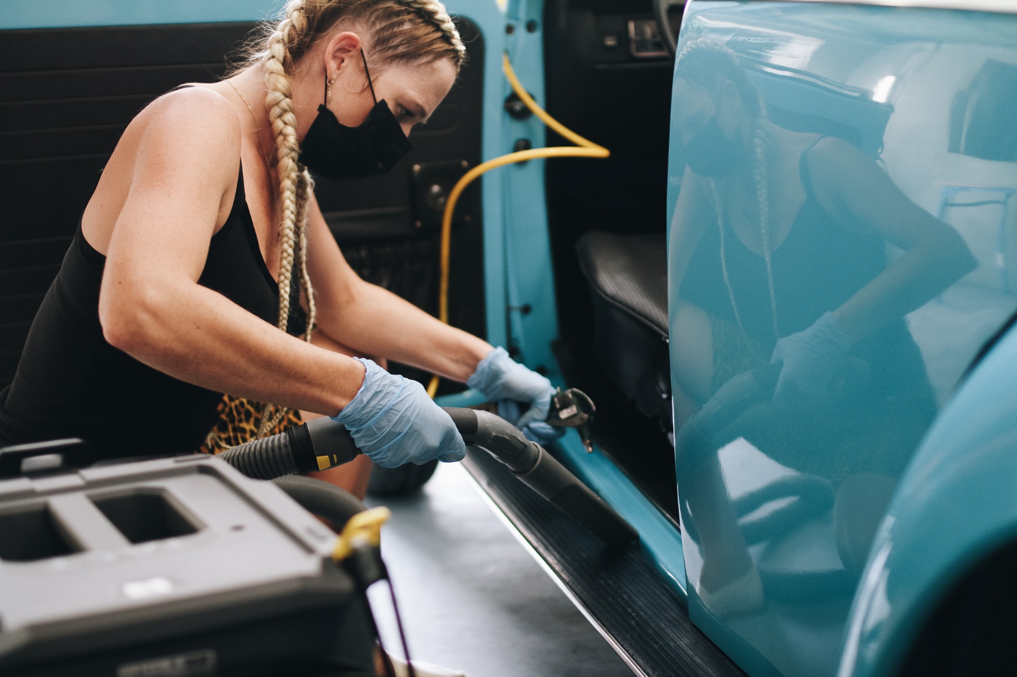 Osoba w masce i rękawicach, z warkoczami, czyści wnętrze turkusowego samochodu odkurzaczem z wężem.
