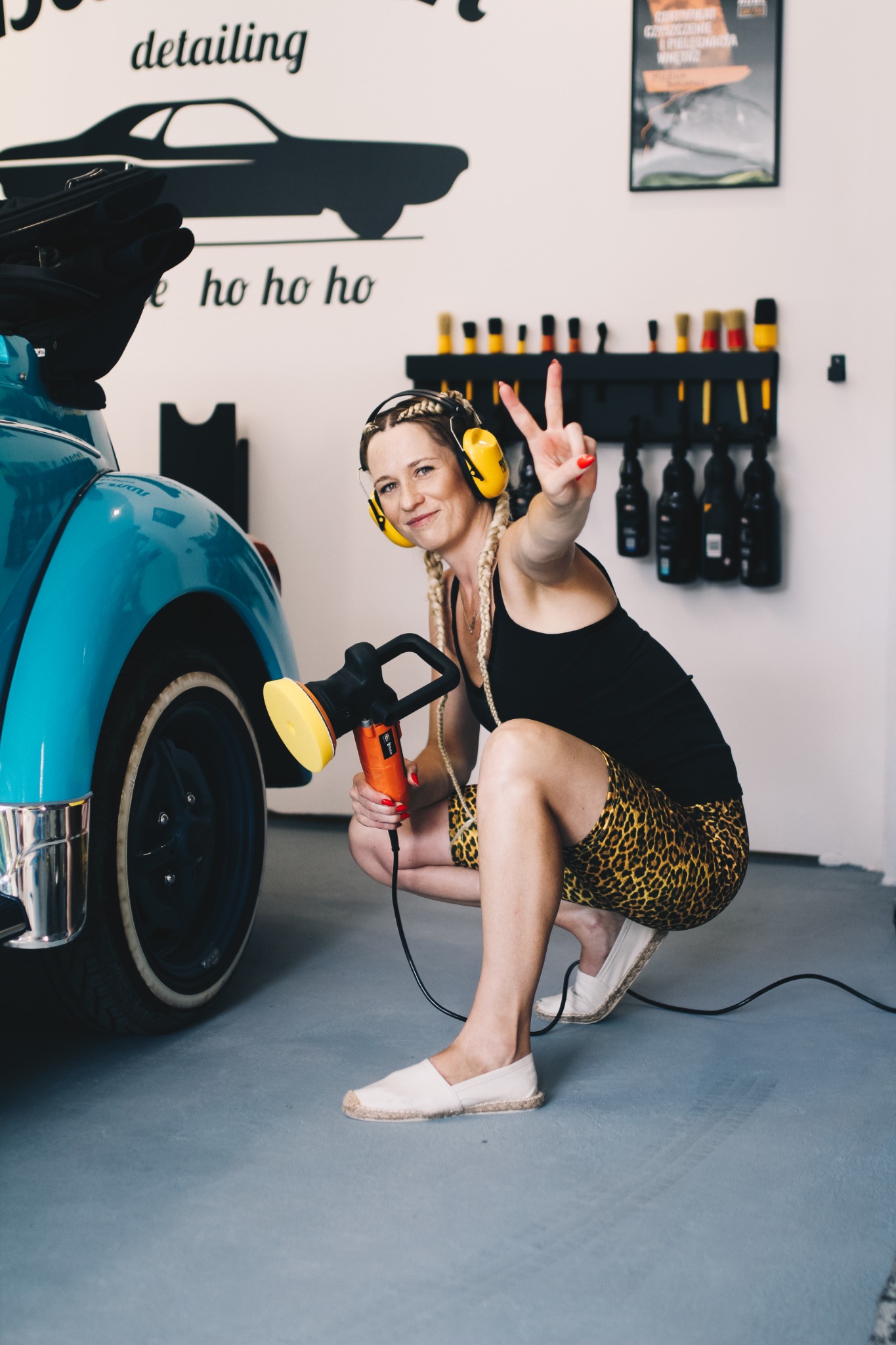 Uśmiechnięta kobieta w słuchawkach i panterkowych spodenkach, kuca obok błękitnego garbusa, trzymając polerkę do lakieru i pokazując znak zwycięstwa, w tle warsztat detailingowy.