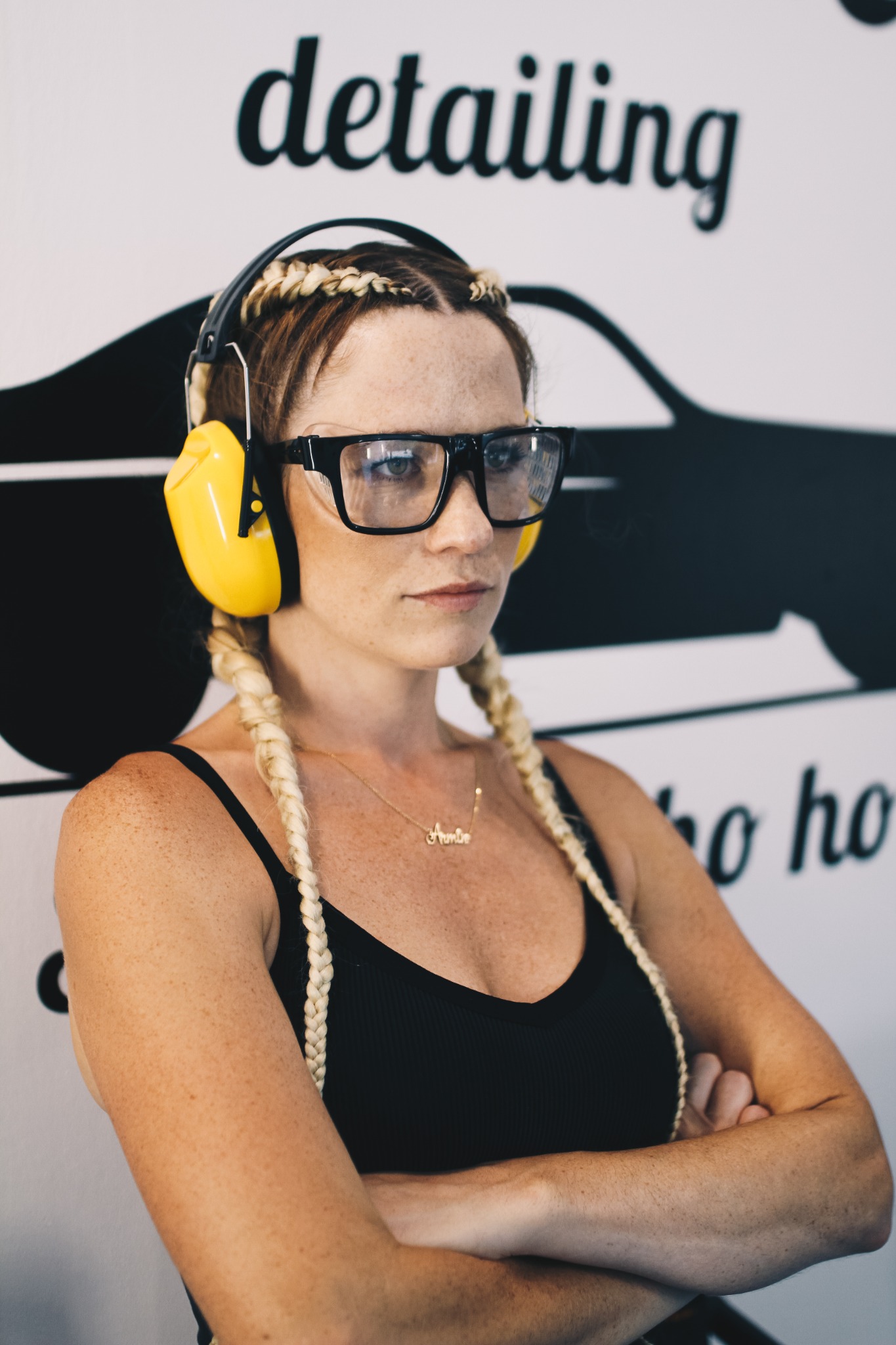 Pracownica firmy detailingowej w Mikołowie, z założonymi słuchawkami ochronnymi i okularami, stoi na tle ściany z grafiką samochodu i napisem 'detailing'.