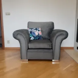 Szary fotel tapicerowany z poduszką w kwiaty, ozdobiony srebrnymi nitami, na drewnianej podłodze.