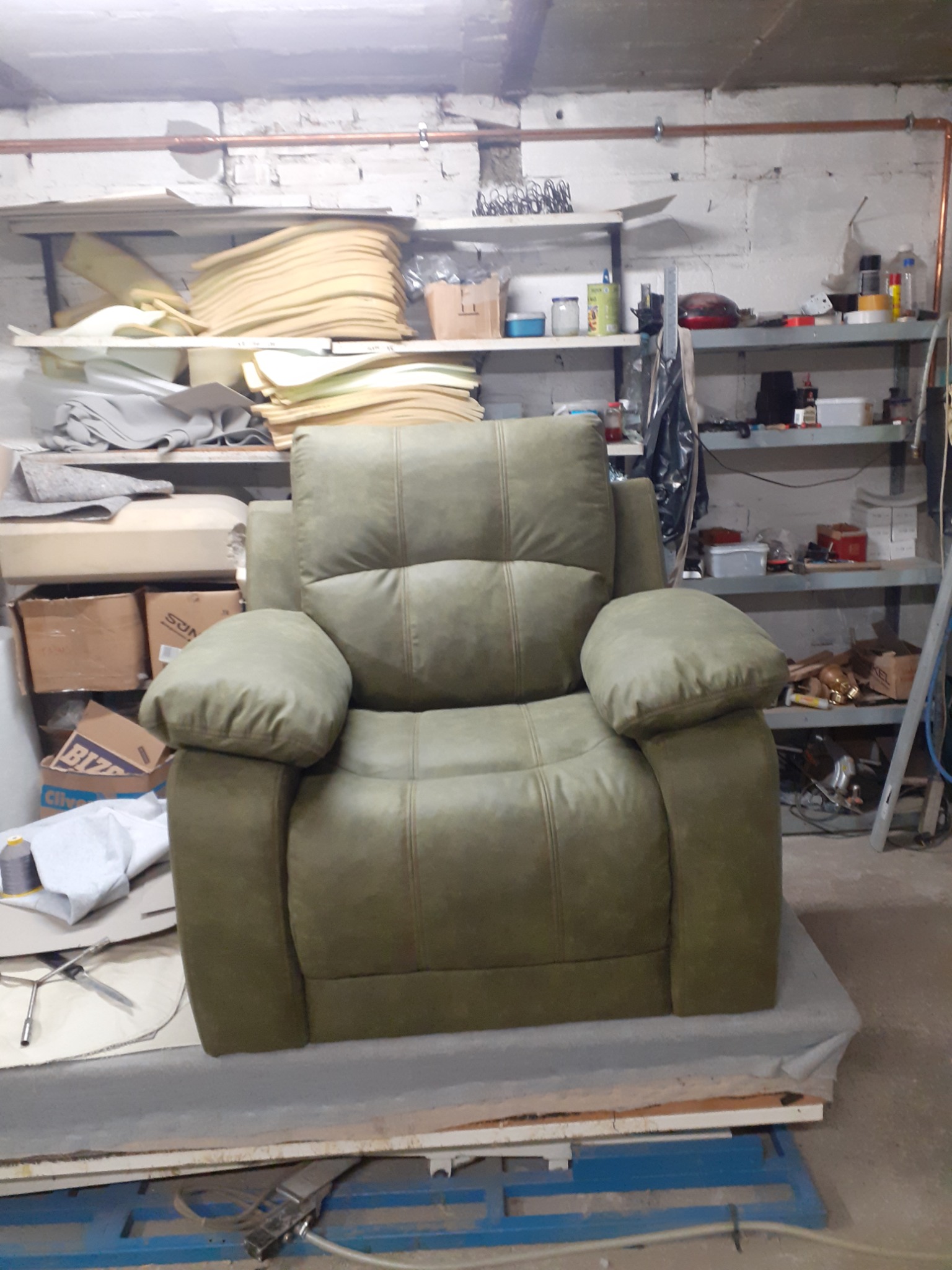 Zielony, tapicerowany fotel z podnoszonym podnóżkiem, stojący w warsztacie tapicerskim; w tle półki z materiałami i narzędziami.