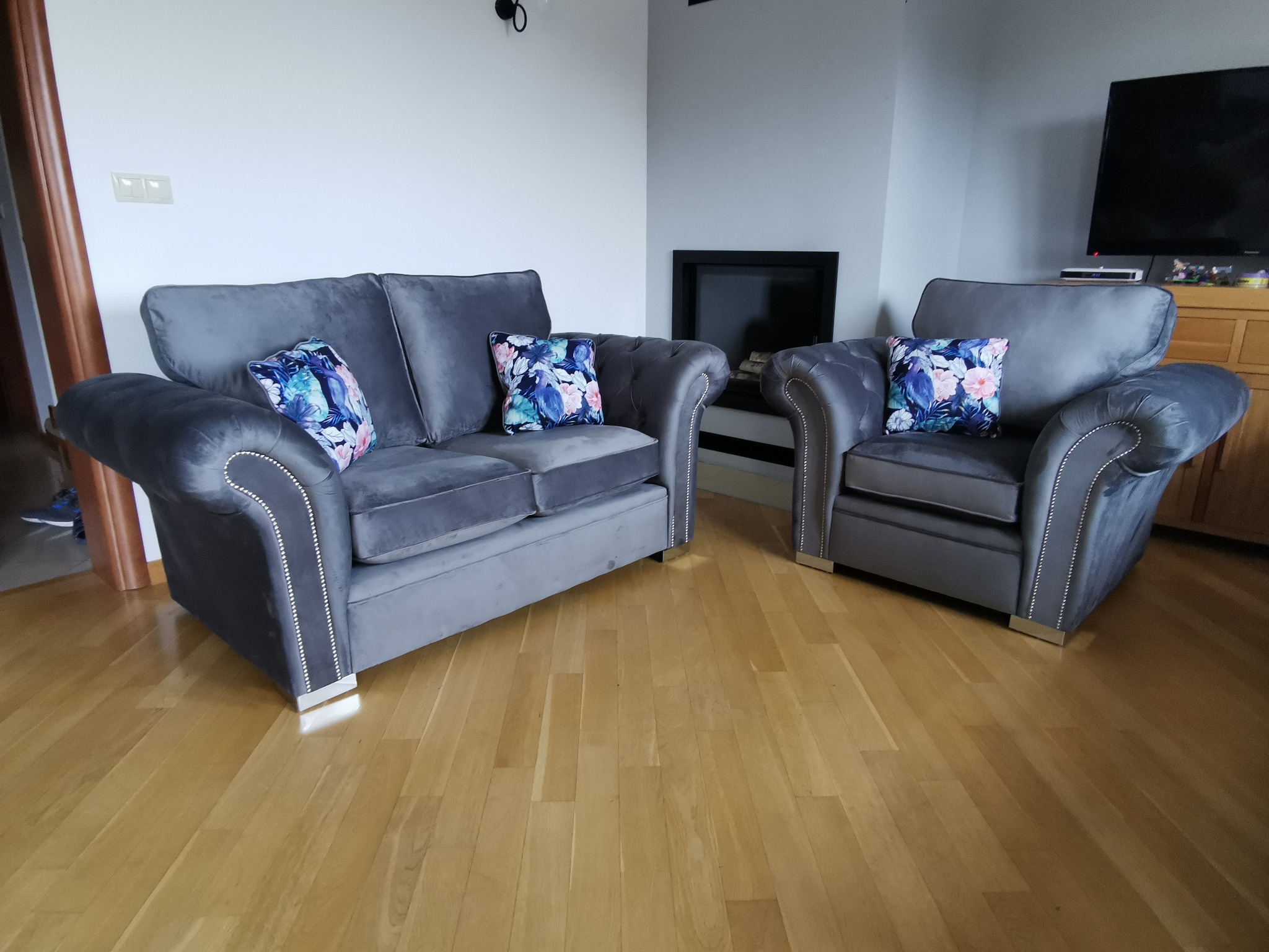 Komplet wypoczynkowy: szara sofa dwuosobowa i fotel z welurową tapicerką, zdobione srebrnymi pinezkami i poduszkami w kwiatowy wzór, na parkiecie.