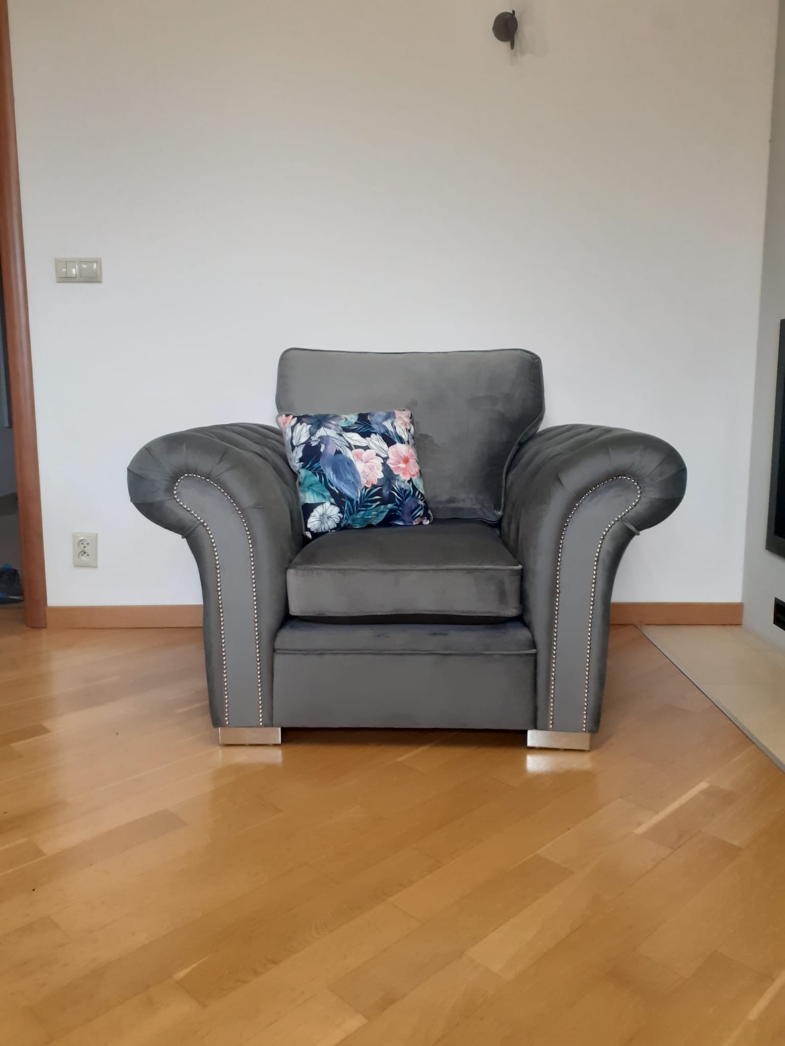Szary fotel tapicerowany z poduszką w kwiaty, ozdobiony srebrnymi nitami, na drewnianej podłodze.