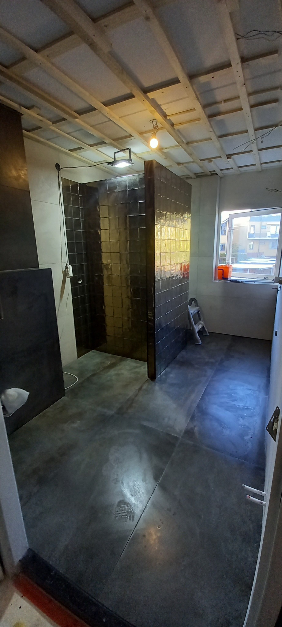 Wnętrze łazienki w trakcie remontu: ciemne, błyszczące płytki w kabinie prysznicowej, surowy beton na podłodze, widoczne elementy instalacji pod sufitem, drabina oparta o ścianę.