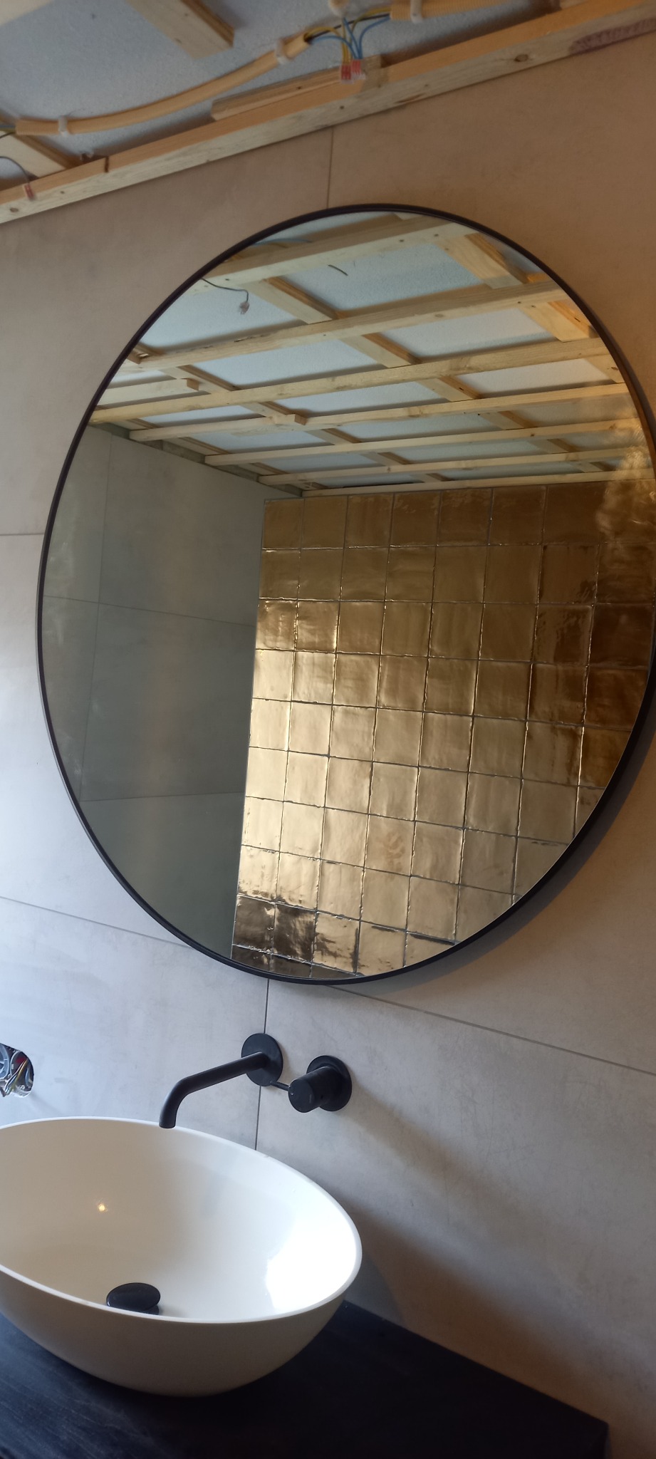 Okrągłe lustro w czarnej ramie odbija ścianę wyłożoną złotymi, kwadratowymi płytkami w łazience w trakcie remontu; widoczna biała umywalka nablatowa i czarna bateria, a w odbiciu w lustrze sufit...