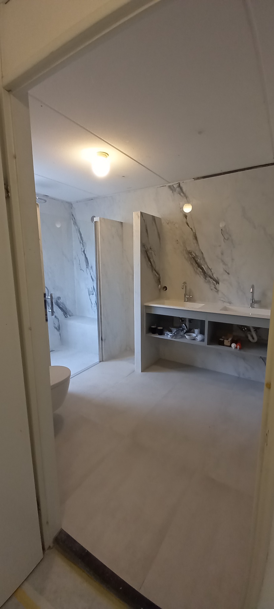 Wykończona łazienka z marmurowymi płytkami na ścianach, kabiną prysznicową i podwójną umywalką z szarą szafką, widziana zza framugi drzwi.