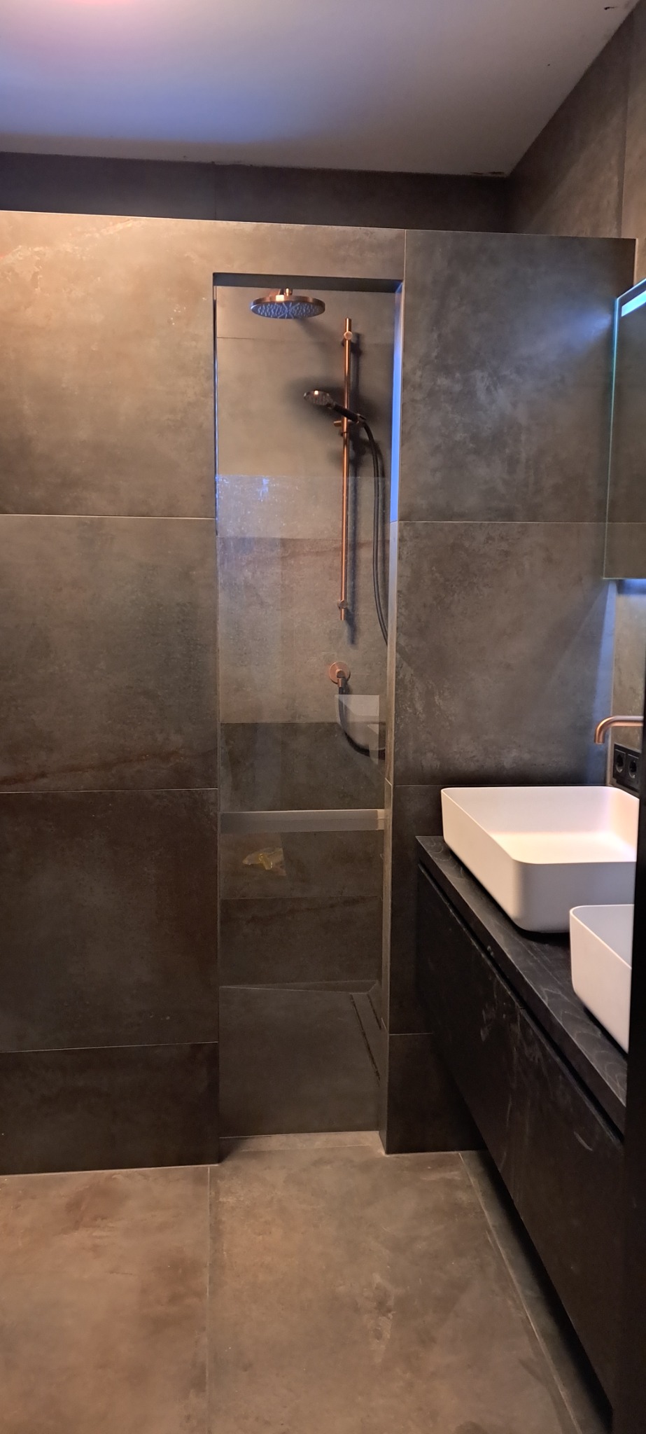 Nowoczesna łazienka z kabiną prysznicową typu walk-in, wykończona płytkami w odcieniach szarości i miedzi, z minimalistyczną umywalką nablatową.