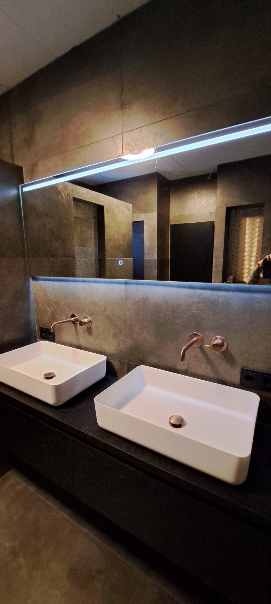 Dwie białe, prostokątne umywalki nablatowe z miedzianymi bateriami na ciemnym blacie; ściany wykończone płytkami imitującymi beton, podświetlane lustro z widocznym odbiciem fragmentu łazienki.