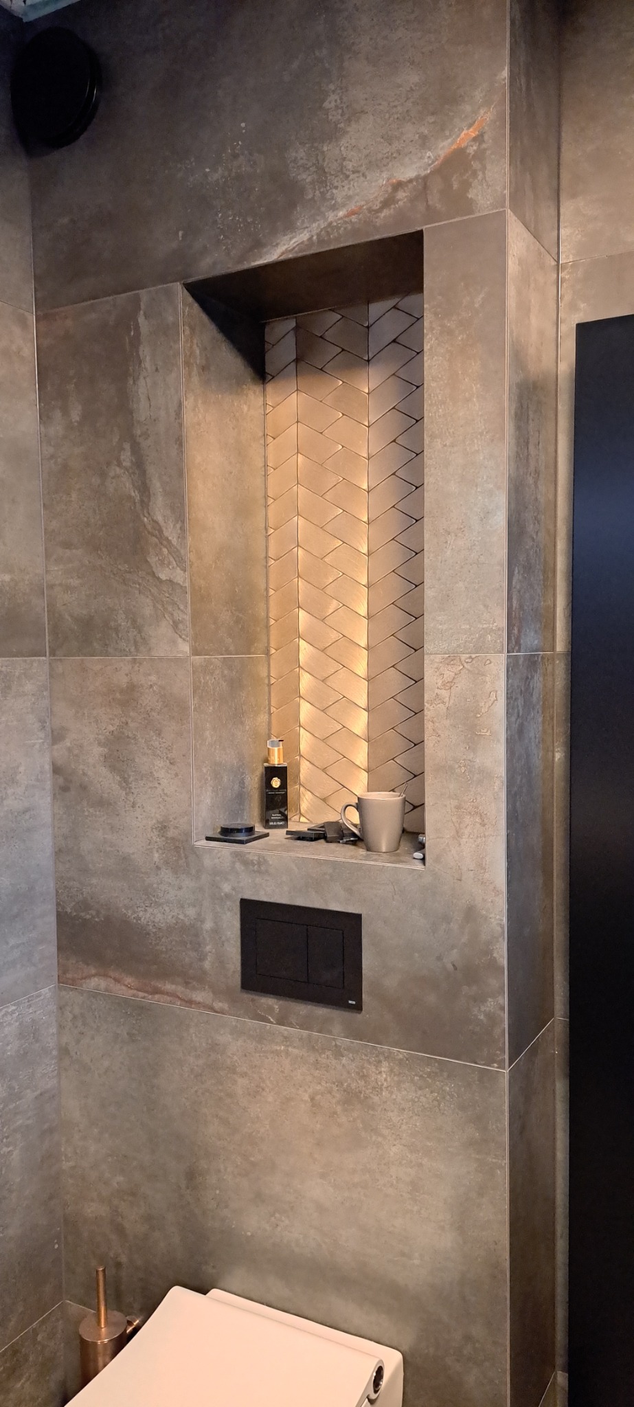 Ściana łazienki wyłożona szarymi płytkami imitującymi kamień, z wnęką wyłożoną złotymi, ułożonymi w jodełkę płytkami, na której stoją flakon perfum i kubek, widoczny fragment toalety.