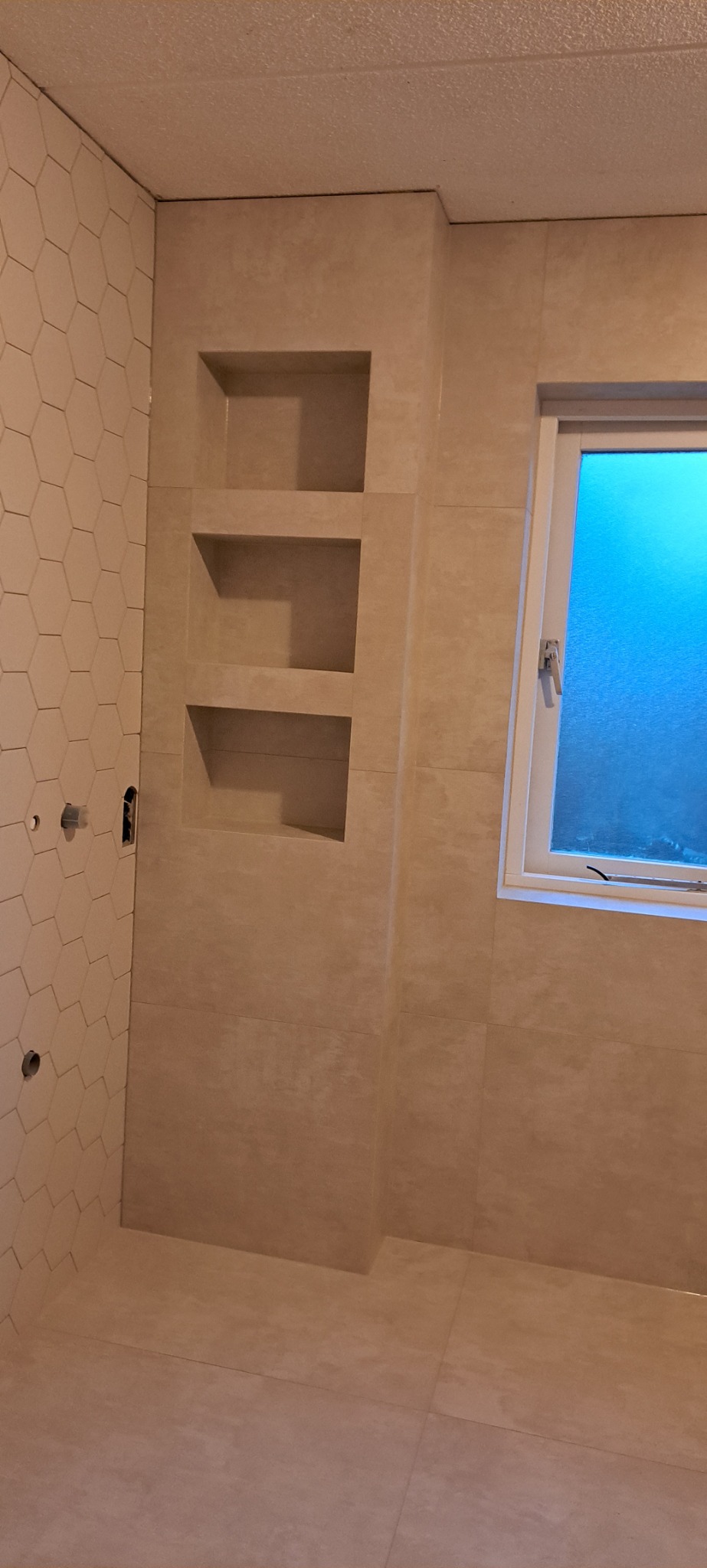 Beżowa łazienka z heksagonalnymi płytkami na jednej ścianie i wnękami w ścianie obok okna, widoczne otwory na armaturę.