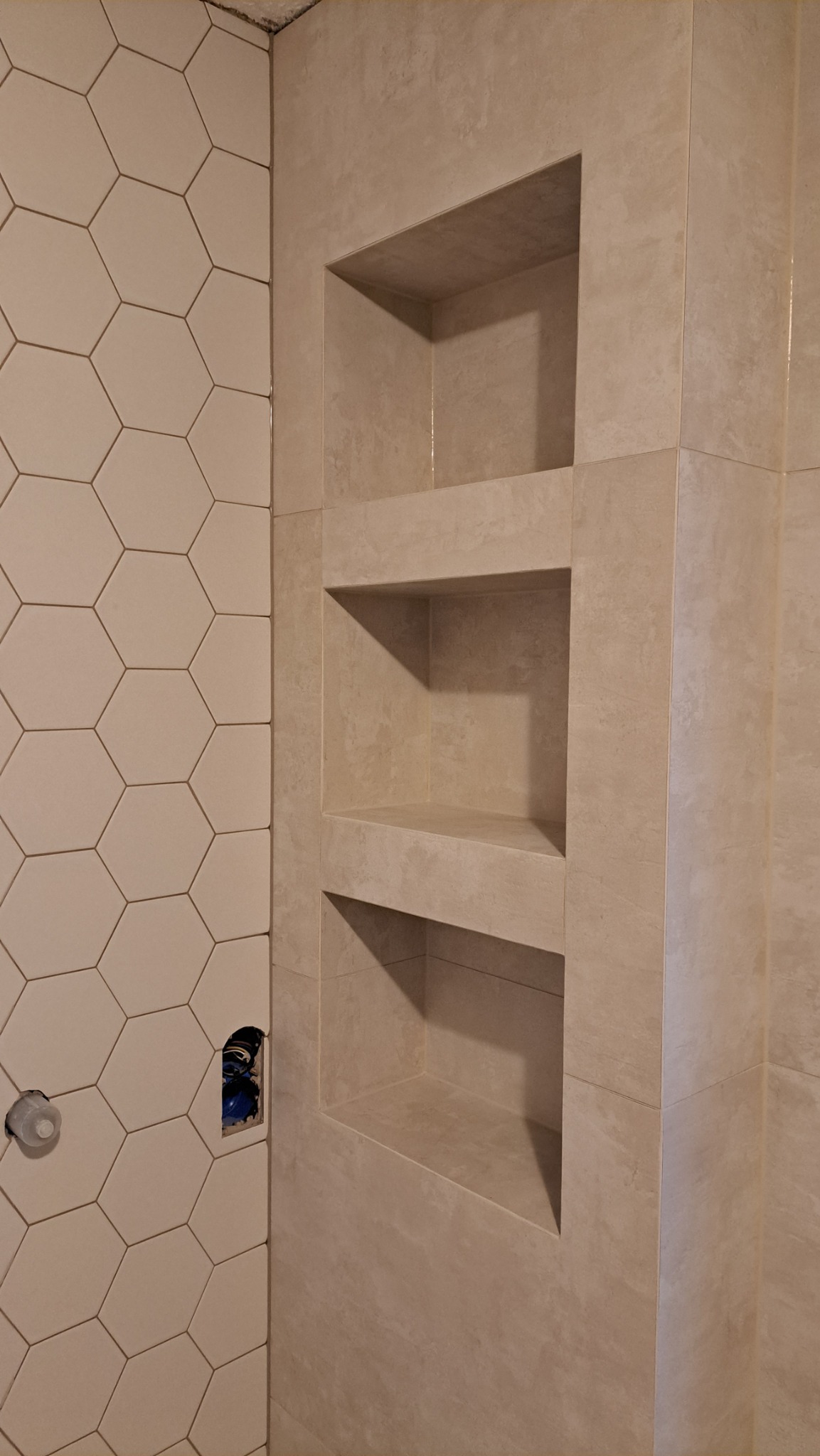 Beżowe heksagonalne płytki na ścianie łazienki sąsiadują z wnękami wyłożonymi płytkami o podobnym odcieniu. Widoczne instalacje pod baterię. Nierówne wykończenie sufitu.