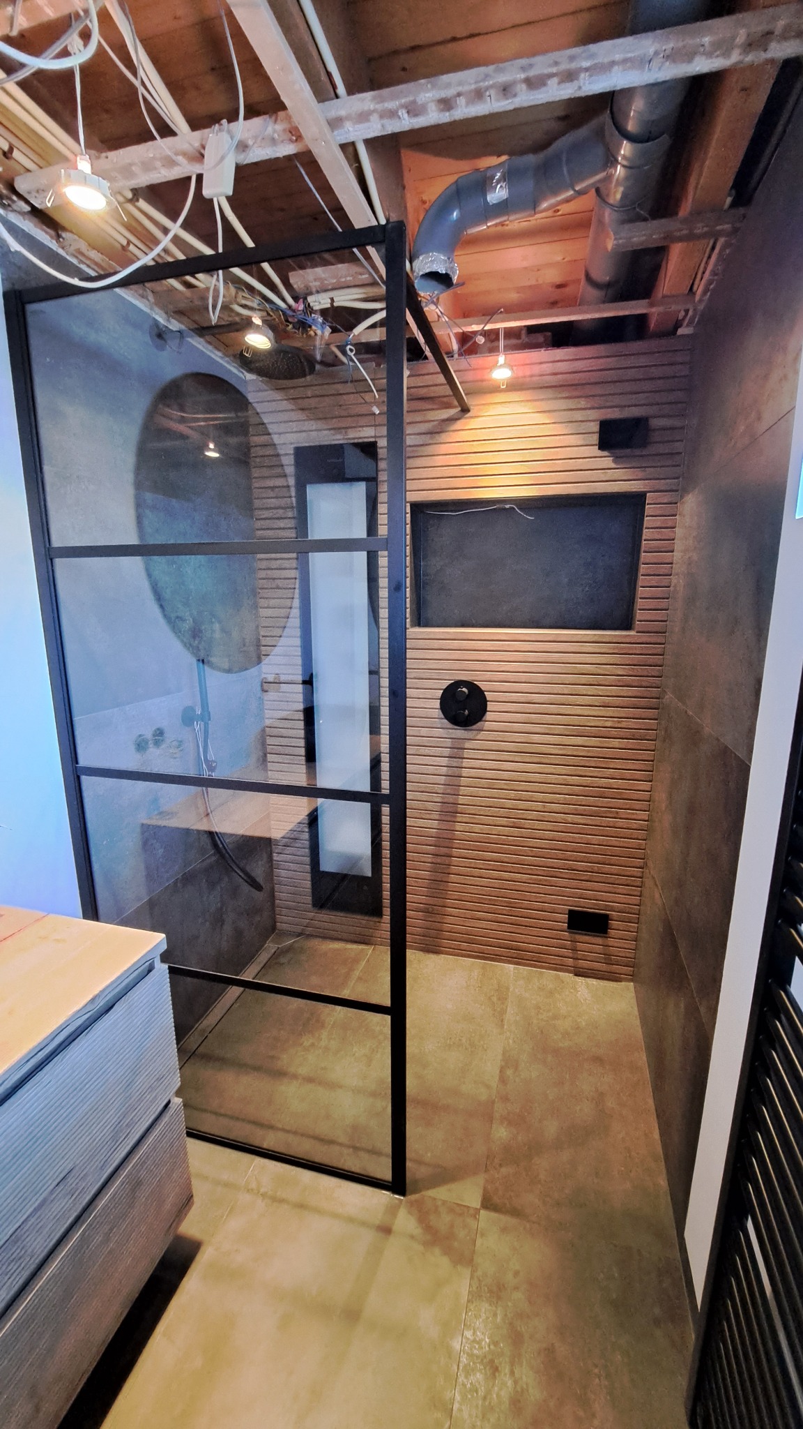 Nowoczesna kabina prysznicowa z czarną ramą, deszczownicą i panelami ściennymi imitującymi drewno, widoczne niedokończone prace na suficie.