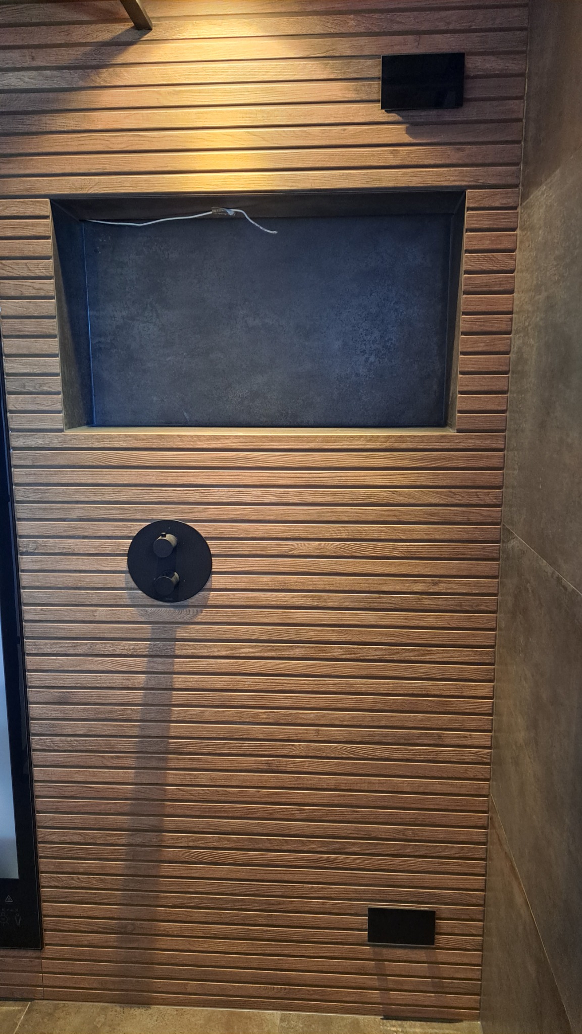 Ściana łazienki wykończona panelami drewnopodobnymi z ukrytym panelem dostępowym i czarną armaturą natryskową.