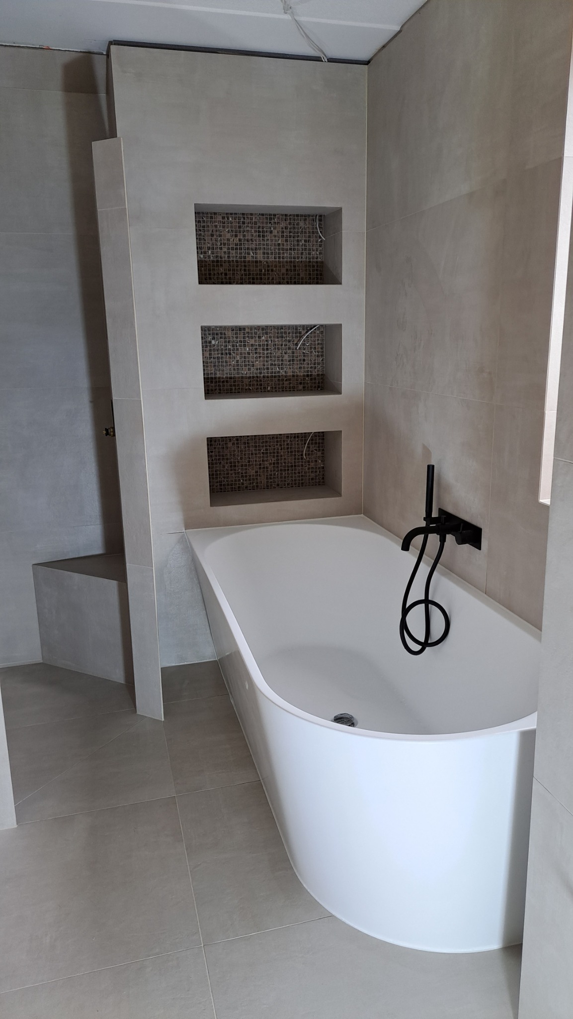 Nowoczesna łazienka w odcieniach szarości z białą wanną i czarną armaturą, trzy wnęki z mozaiką nad wanną, widoczne przewody elektryczne w wnękach.