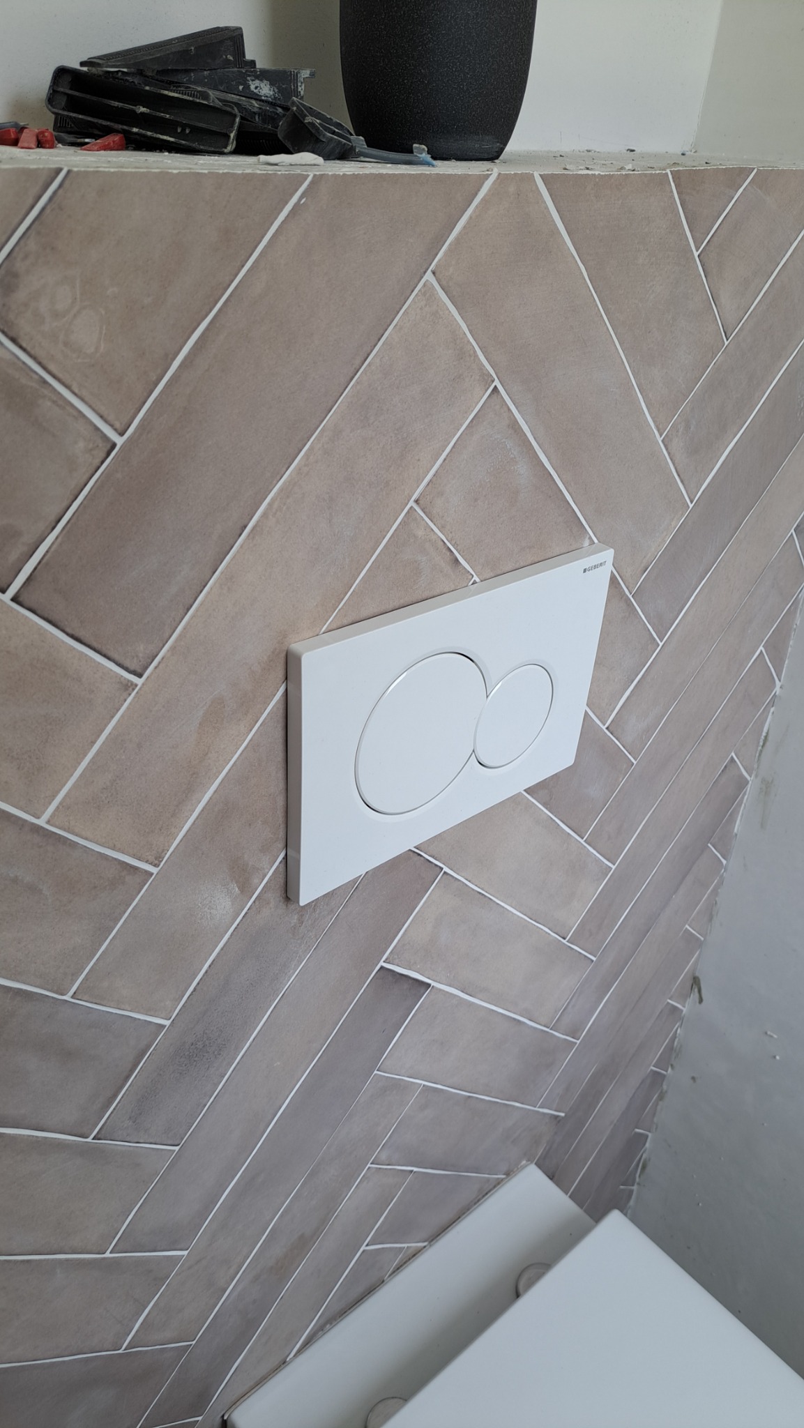 Beżowe płytki ułożone w jodełkę na ścianie w łazience, z zamontowanym białym panelem sterującym spłuczką Geberit i widocznymi narzędziami glazurniczymi na górze.