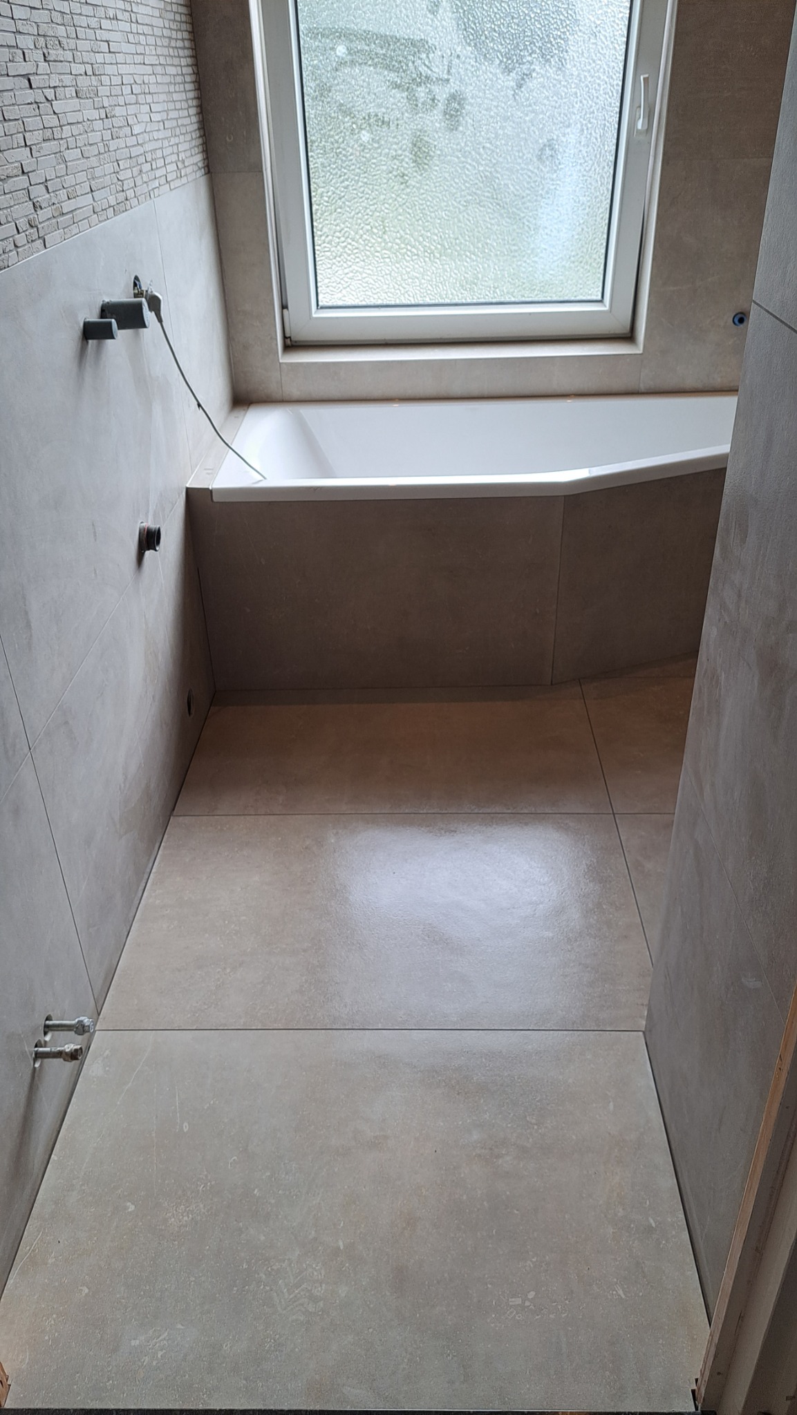 Wykończona łazienka z beżowymi, wielkoformatowymi płytkami na podłodze i ścianach, białą wanną pod oknem dachowym i wystającymi elementami instalacji wodnej.