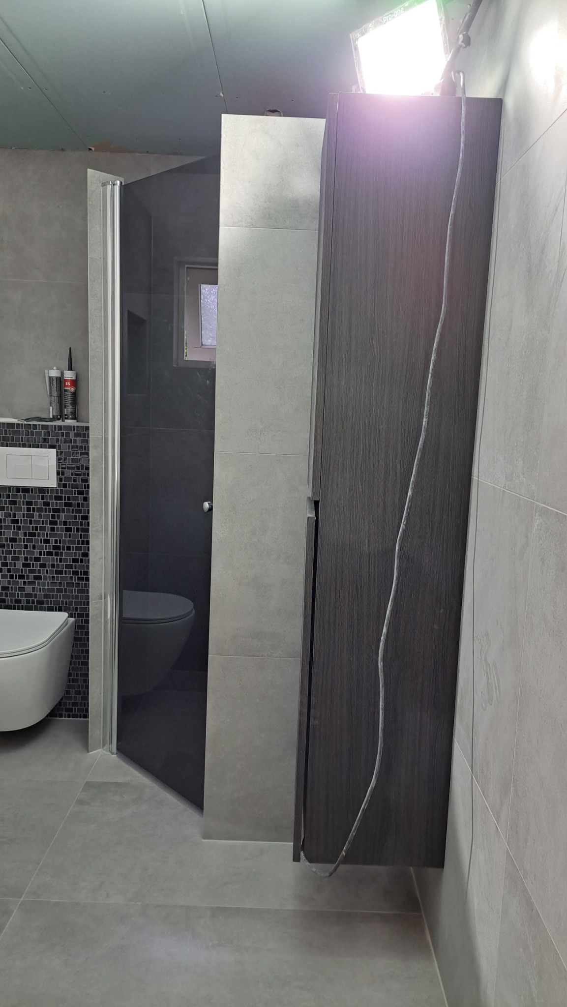 Szara łazienka w trakcie wykańczania: widoczna kabina prysznicowa z ciemnymi płytkami, wisząca toaleta, mozaika nad stelażem, szafka z ciemnego drewna oraz lampa budowlana oświetlająca pomieszczenie.