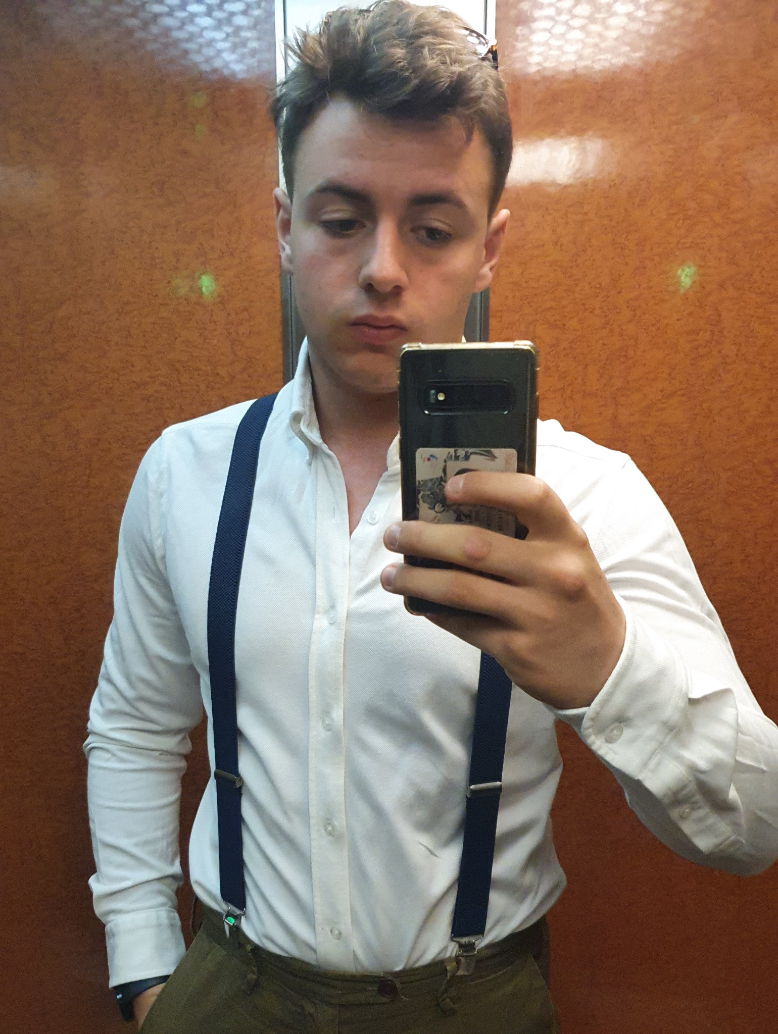 Młody mężczyzna w białej koszuli i granatowych szelkach robi selfie w windzie, trzymając czarny telefon z naklejką na obudowie.