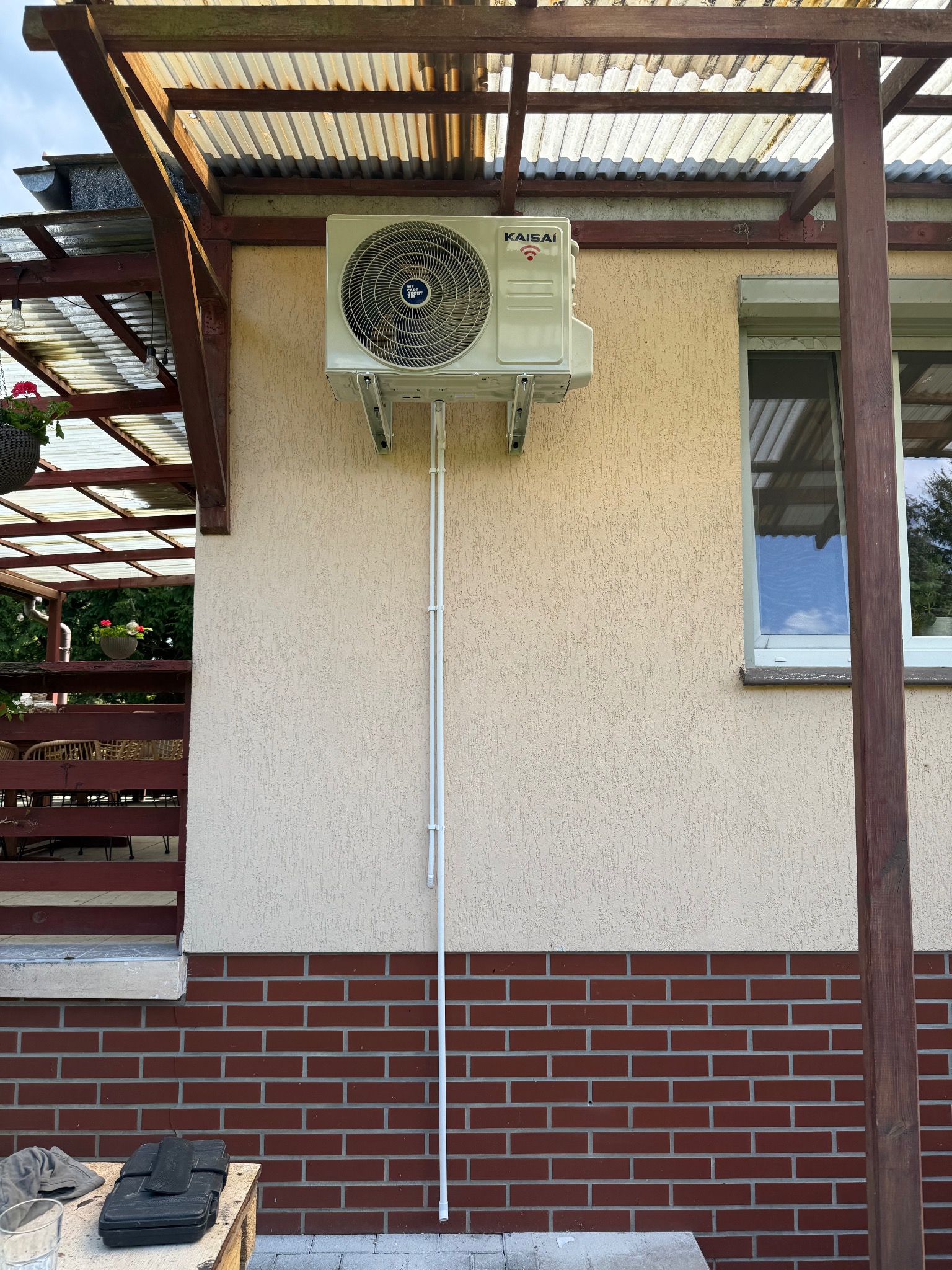 Zamontowana jednostka zewnętrzna klimatyzacji Kaisai na elewacji budynku z brązową cegłą i drewnianą konstrukcją zadaszenia. Białe rurki odprowadzające.