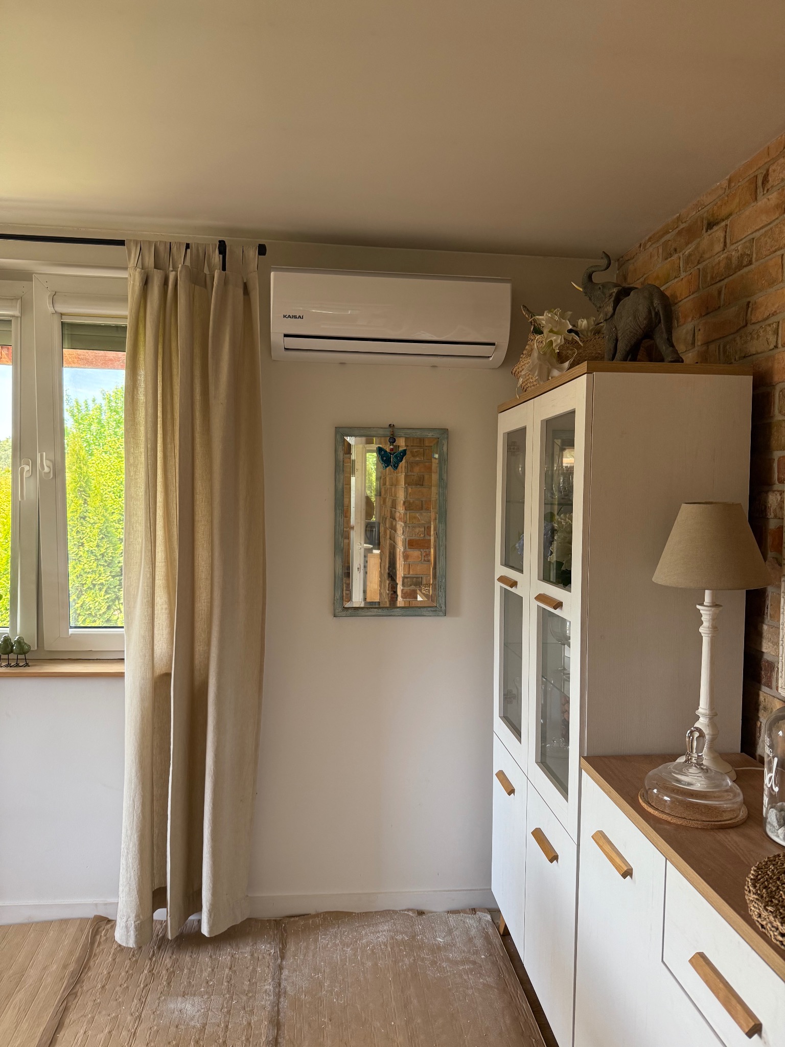 Biała klimatyzacja Kaisai zamontowana w pokoju z ceglaną ścianą, zasłoniętym oknem i białą komodą. Widoczne zabezpieczone podłogi podczas montażu.