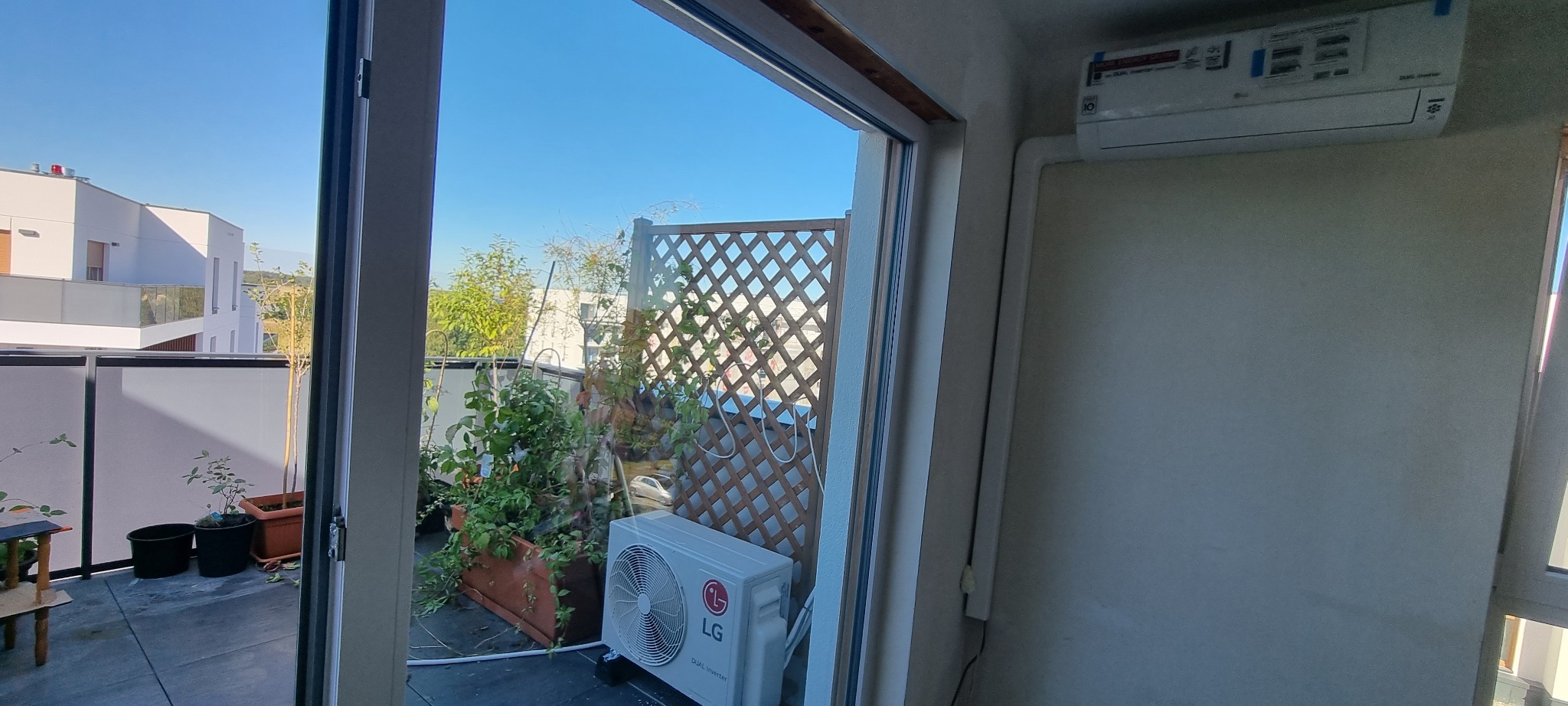 Widok z mieszkania na balkon z jednostką zewnętrzną klimatyzacji LG Dual Inverter, kratką osłonową i roślinami w donicach, na ścianie wewnątrz zamontowana jednostka wewnętrzna.