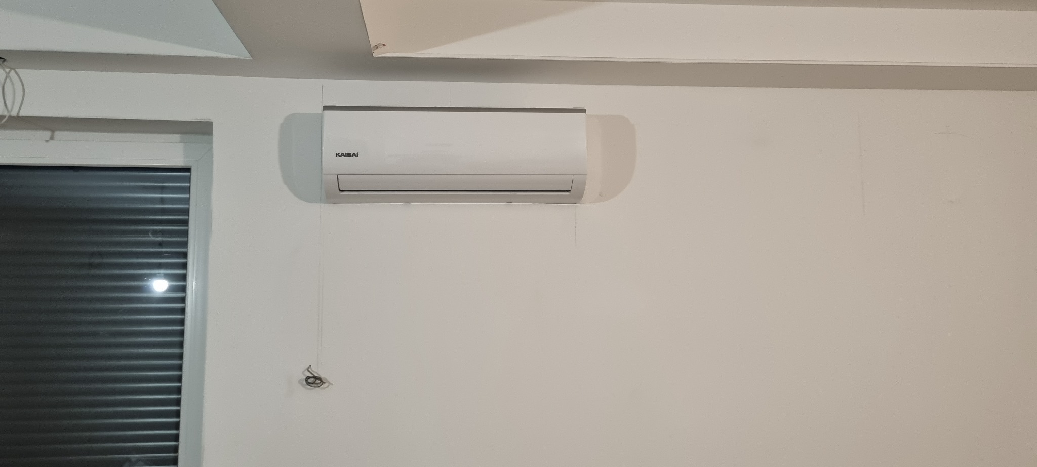Biały klimatyzator Kaisai zamontowany na białej ścianie w pomieszczeniu z żaluzjami w oknie. Widoczne wystające kable.
